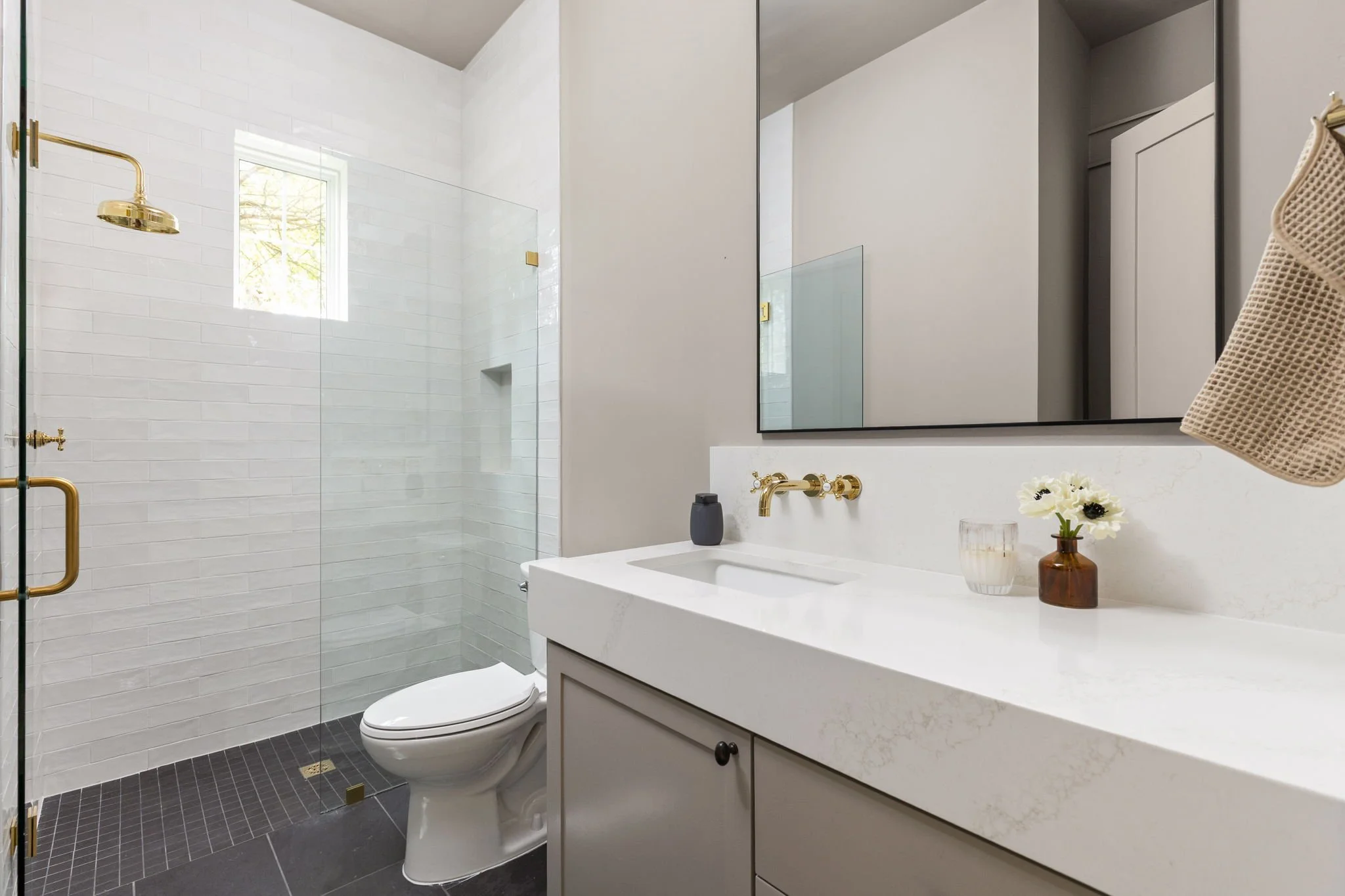 Modern bathroom featuring a walk-in shower with glass door, a white sink and vanity with decorative flowers, a large mirror, gold fixtures, and dark tiled flooring.