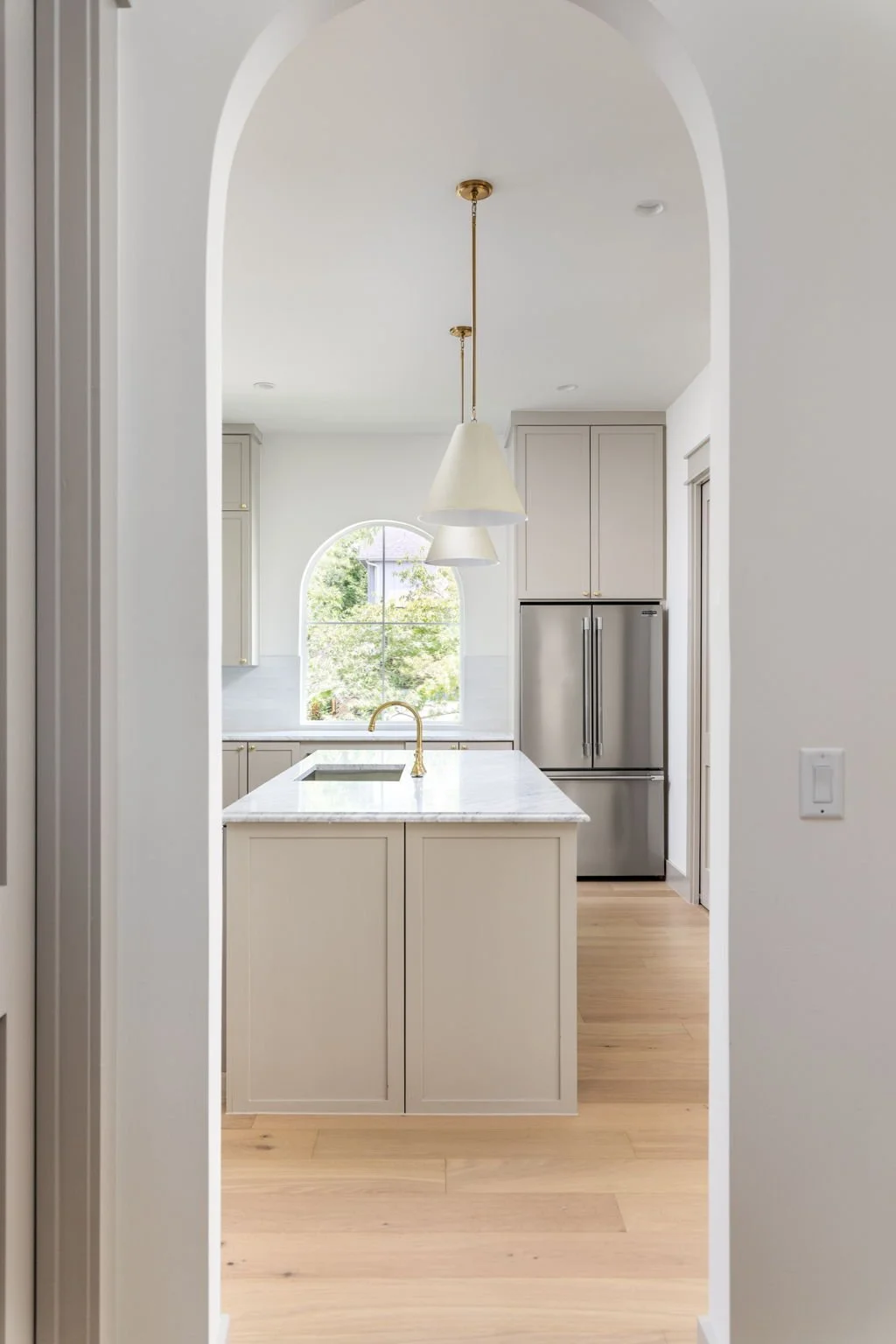 Modern kitchen with white cabinets, marble countertop, gold faucet, stainless steel refrigerator, and an arched window with greenery outside, seen through an opening in a white wall.
