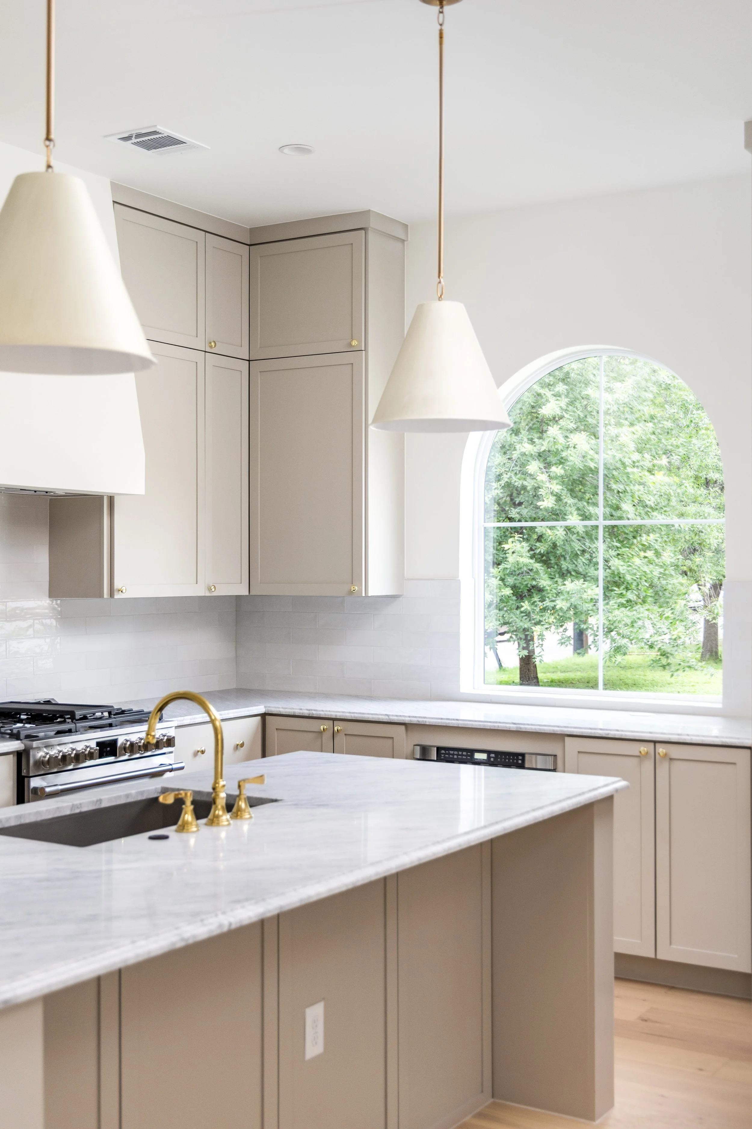 Kitchen with beige cabinetry, white marble countertops, a window showing green trees outside, and two white pendant lights hanging from the ceiling.