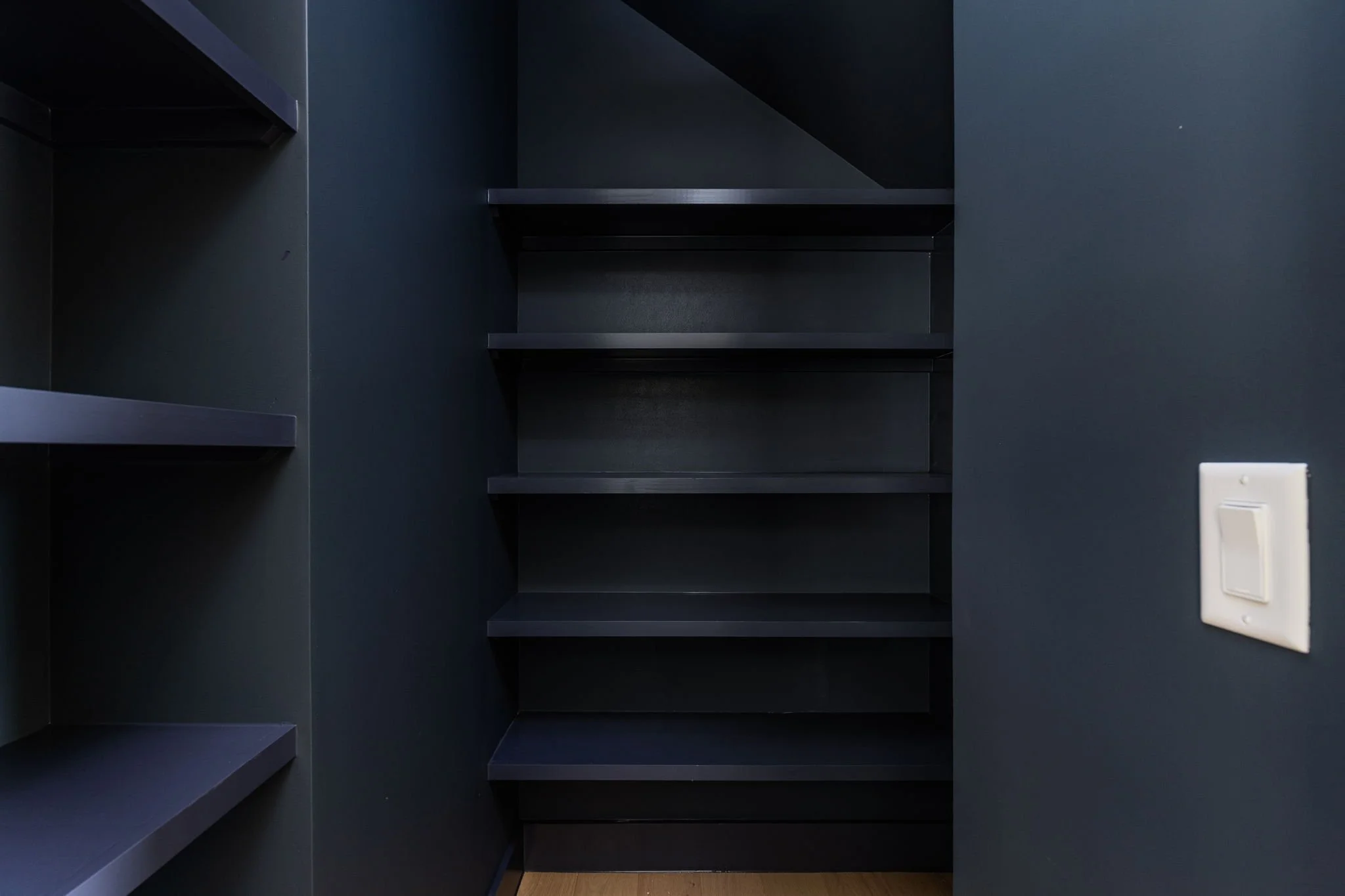 Empty black shelving unit with five shelves against a dark wall, next to a light switch on the right.
