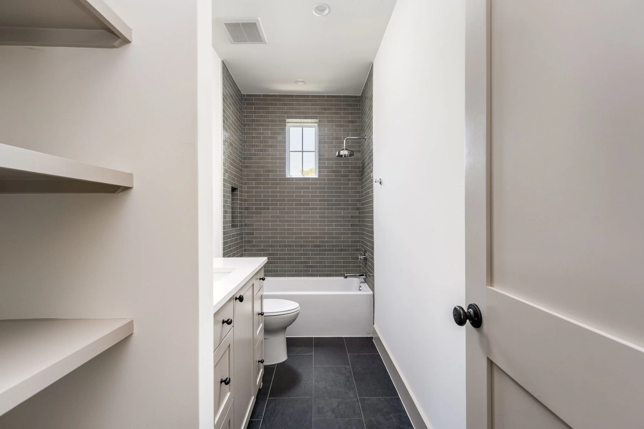 Bathroom with a gray tiled shower, white vanity, toilet, small window, and white walls.