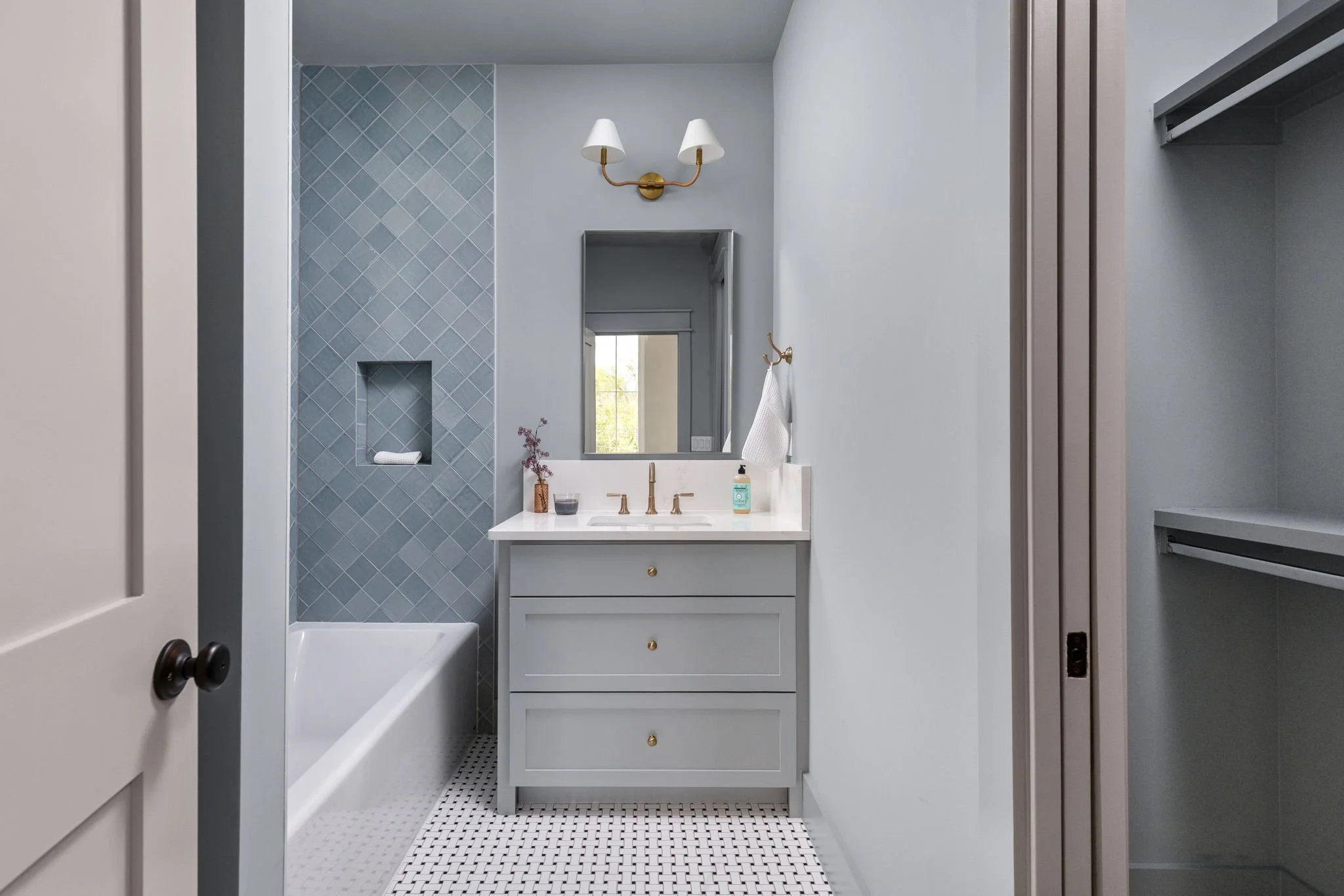 View of a modern bathroom vanity with a white sink, gold fixtures, and a mirror. Blue tile shower with ledge and niche, and a window showing greenery outside. The floor has small black-and-white tile, and the walls are painted a light gray.