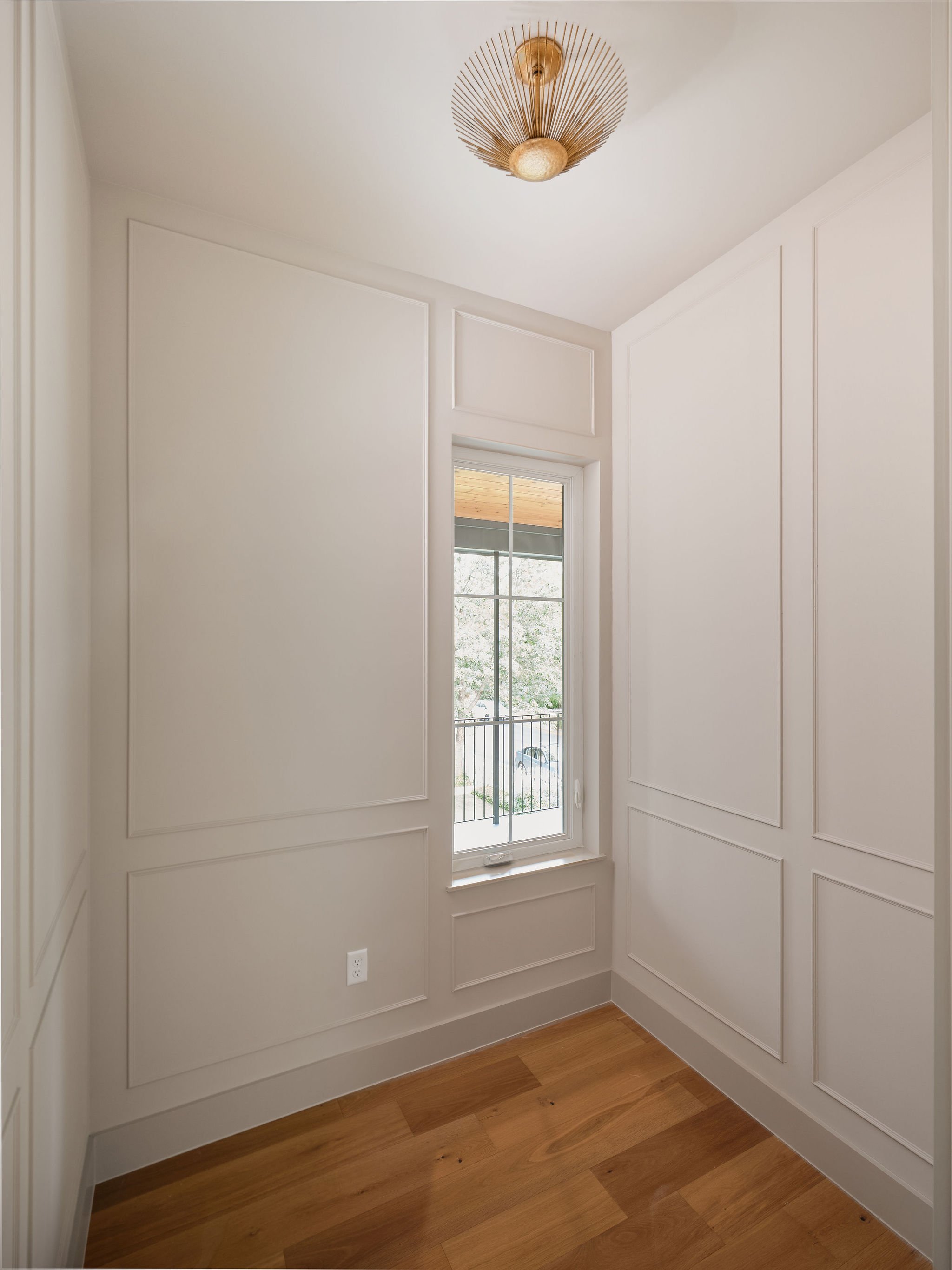 Small room with white paneled walls, a window with a view of a porch and trees, a wood floor, and a ceiling light fixture.