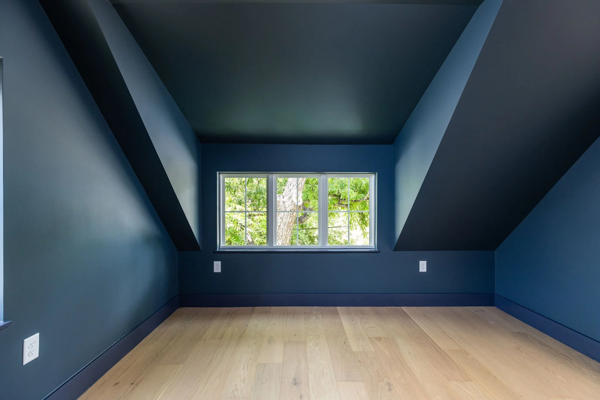 Empty room with blue walls and angled ceiling, featuring a large window showing greenery outside, with hardwood floor and electrical outlets.