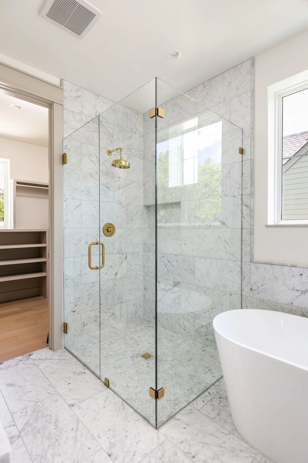 Modern bathroom featuring a glass shower with brass fixtures, a white freestanding bathtub, marble tiles, and a window showing outdoor trees.