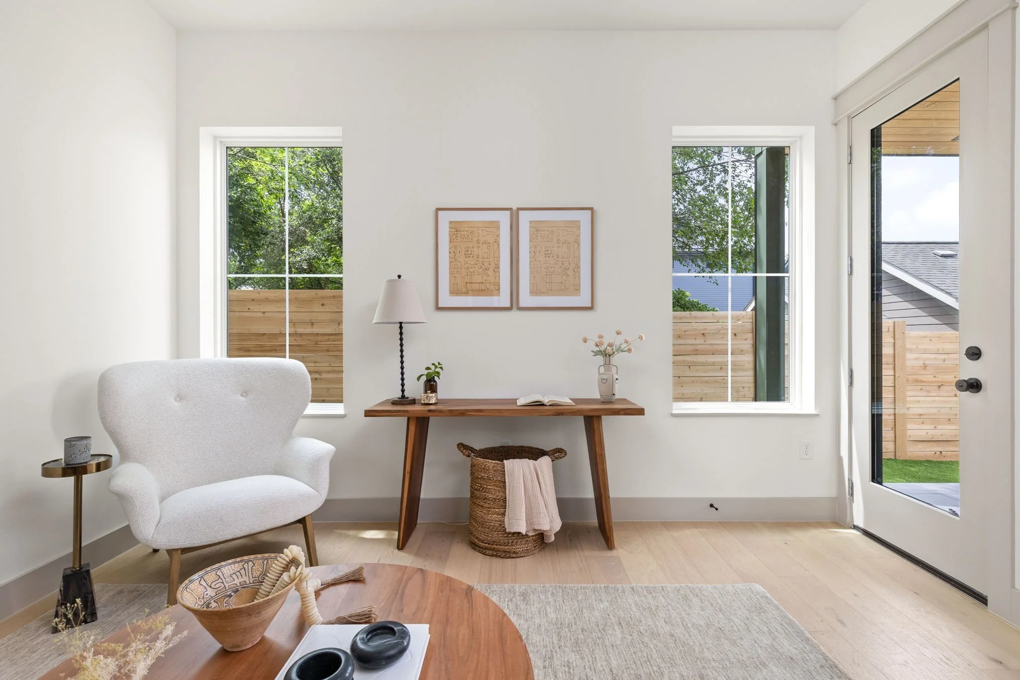 A bright living room with white walls and large windows showing a backyard with green trees and a wooden fence. There is a white upholstered armchair to the left, a small side table with a candle, and a round wooden coffee table with decorative bowls