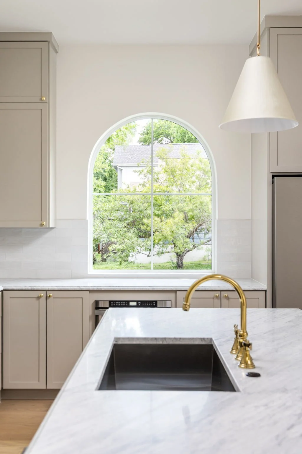 View of a kitchen with beige cabinets, white marble countertops, a black under-mount sink with a gold faucet, and a large arched window revealing a green backyard with trees.