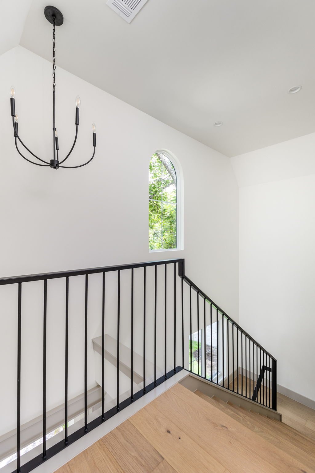 Interior view of a staircase landing with a black metal railing, a large arched window with greenery outside, white walls, hardwood flooring, and a black chandelier hanging from the ceiling.