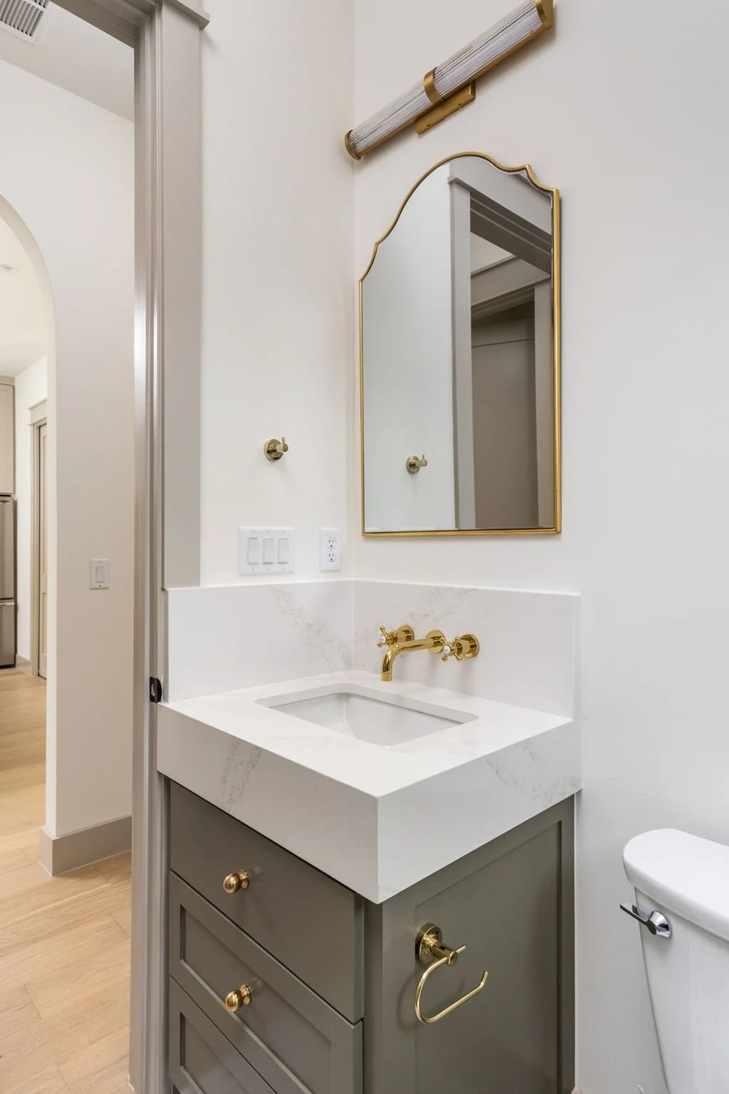 Bathroom vanity with a small white sink, gold fixtures, a gray cabinet with drawers, and a rectangular mirror with a gold frame. A light fixture is mounted above the mirror, and a white toilet is partially visible on the right.