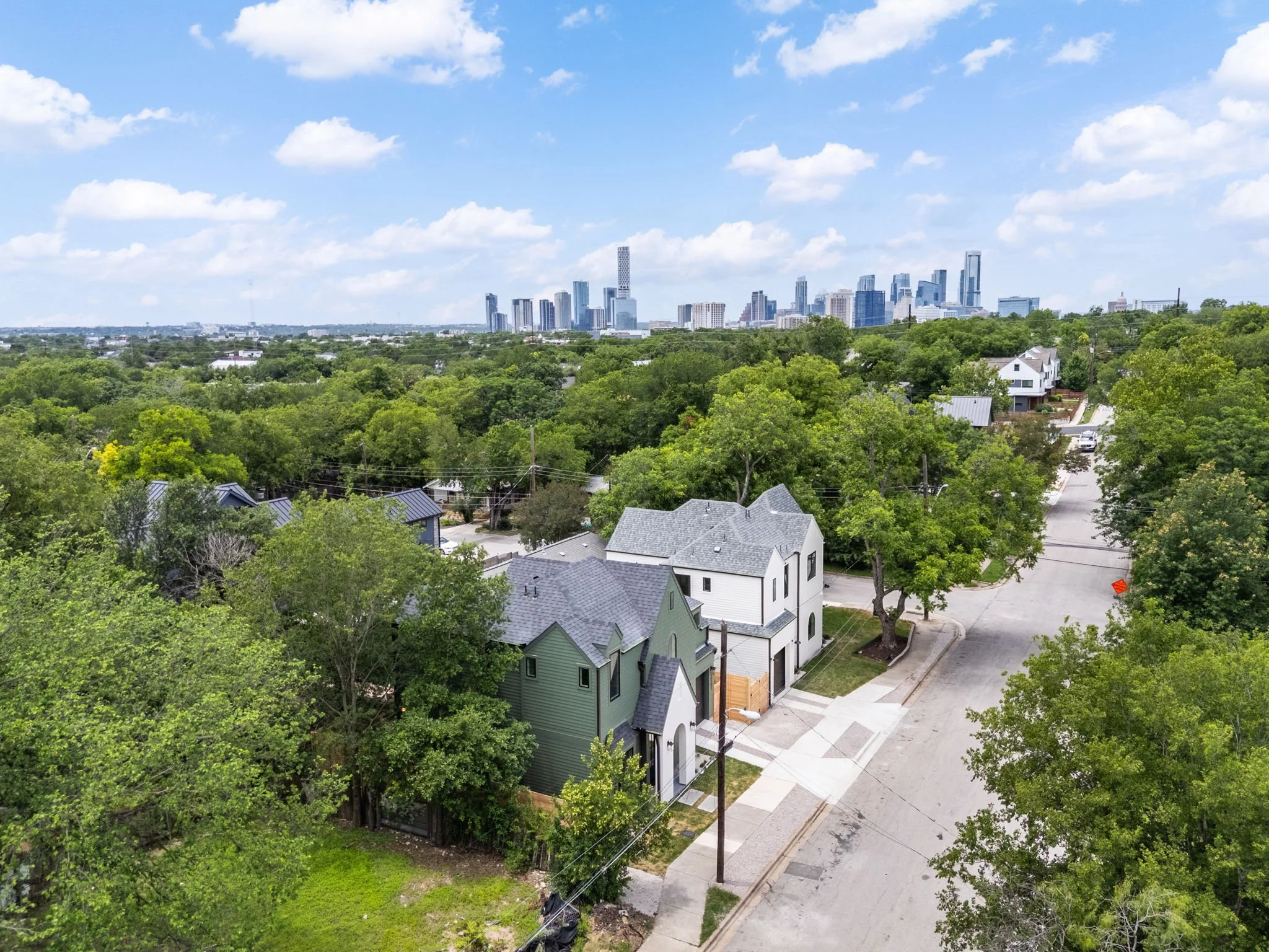 A suburban neighborhood with modern houses, green trees, and a city skyline in the background under a partly cloudy sky.