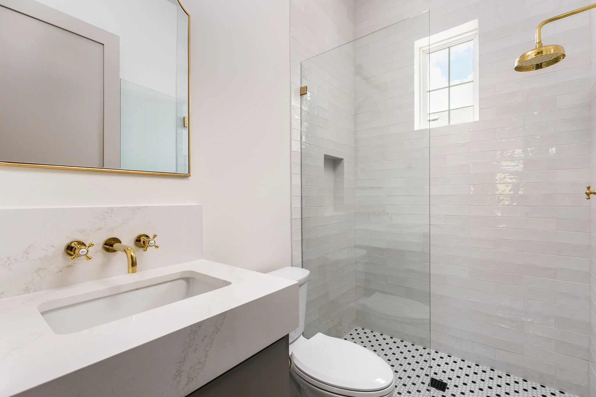 Modern bathroom with white marble vanity, gold fixtures, and walk-in shower with white subway tiles and a window.