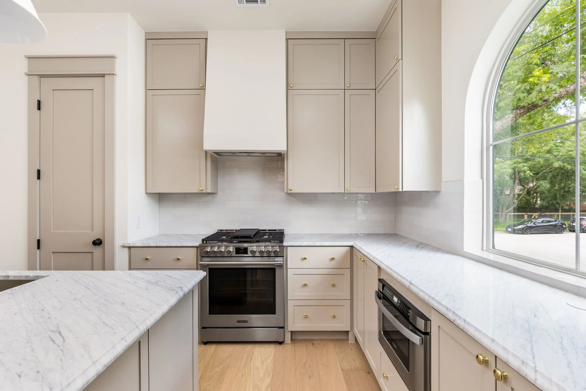 A modern kitchen with beige cabinets, white marble countertops, stainless steel oven, and a large arched window showing green trees outside.