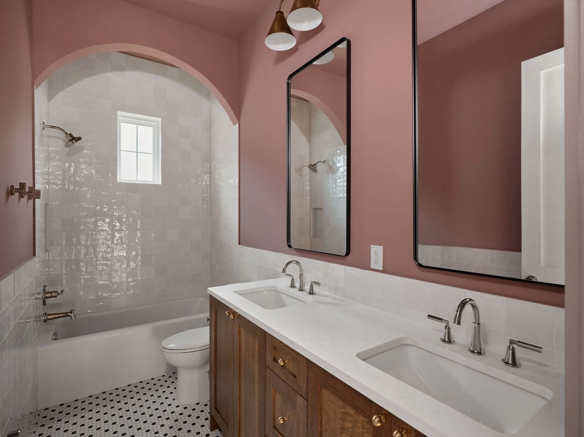 Bathroom with pink walls, double vanity with marble countertop and two sinks, two mirrors, wooden cabinet, bathtub with shower, window, small tile floor.