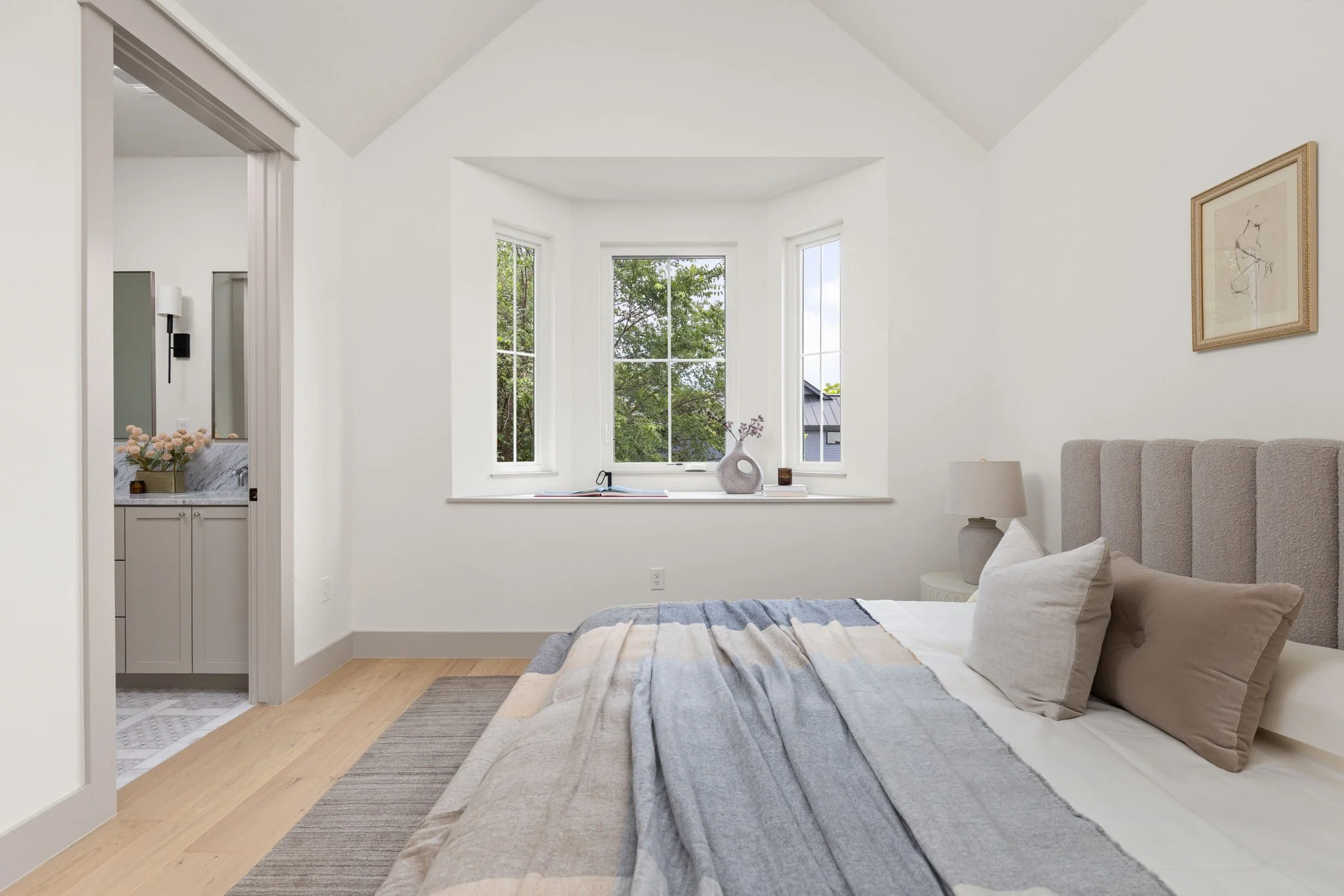 A bedroom with white walls, a beige upholstered bed, a gray and beige striped blanket, and matching pillows. Large windows show green trees outside. Beside the bed is a nightstand with a white lamp. A decorative vase and books are on the window ledge