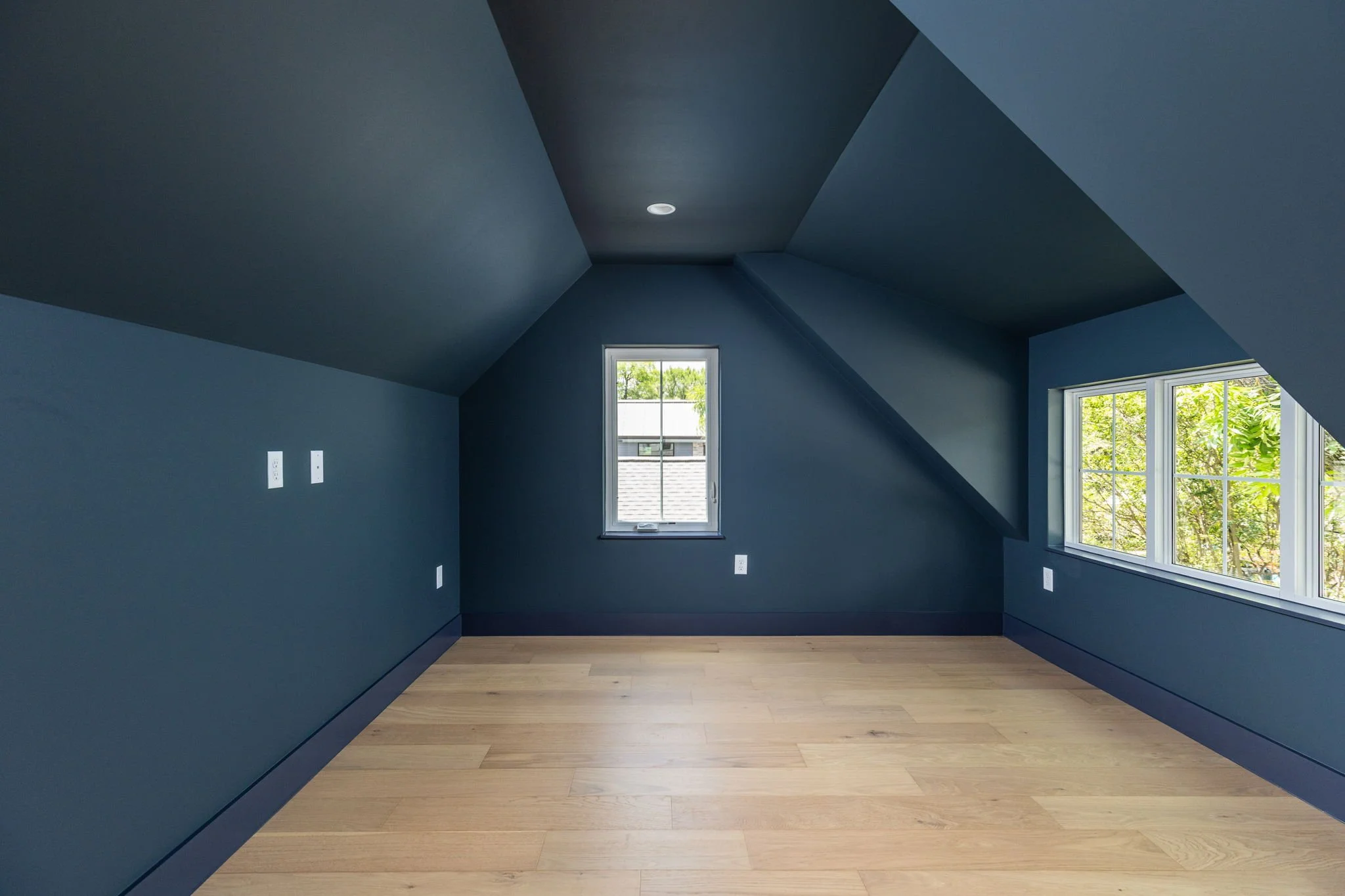 Empty attic room with blue walls and ceiling, two windows showing green trees outside, and light wooden flooring.