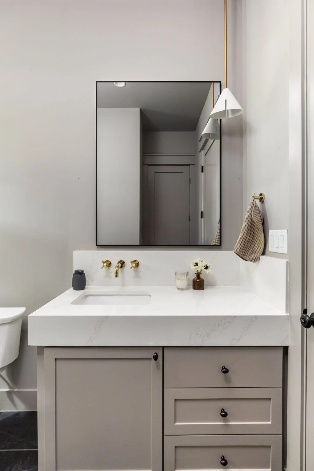 Modern bathroom with a gray cabinet vanity featuring a white marble countertop, gold fixtures, a large mirror, and decor including a small vase with white flowers, a candle, and a gray soap dispenser.