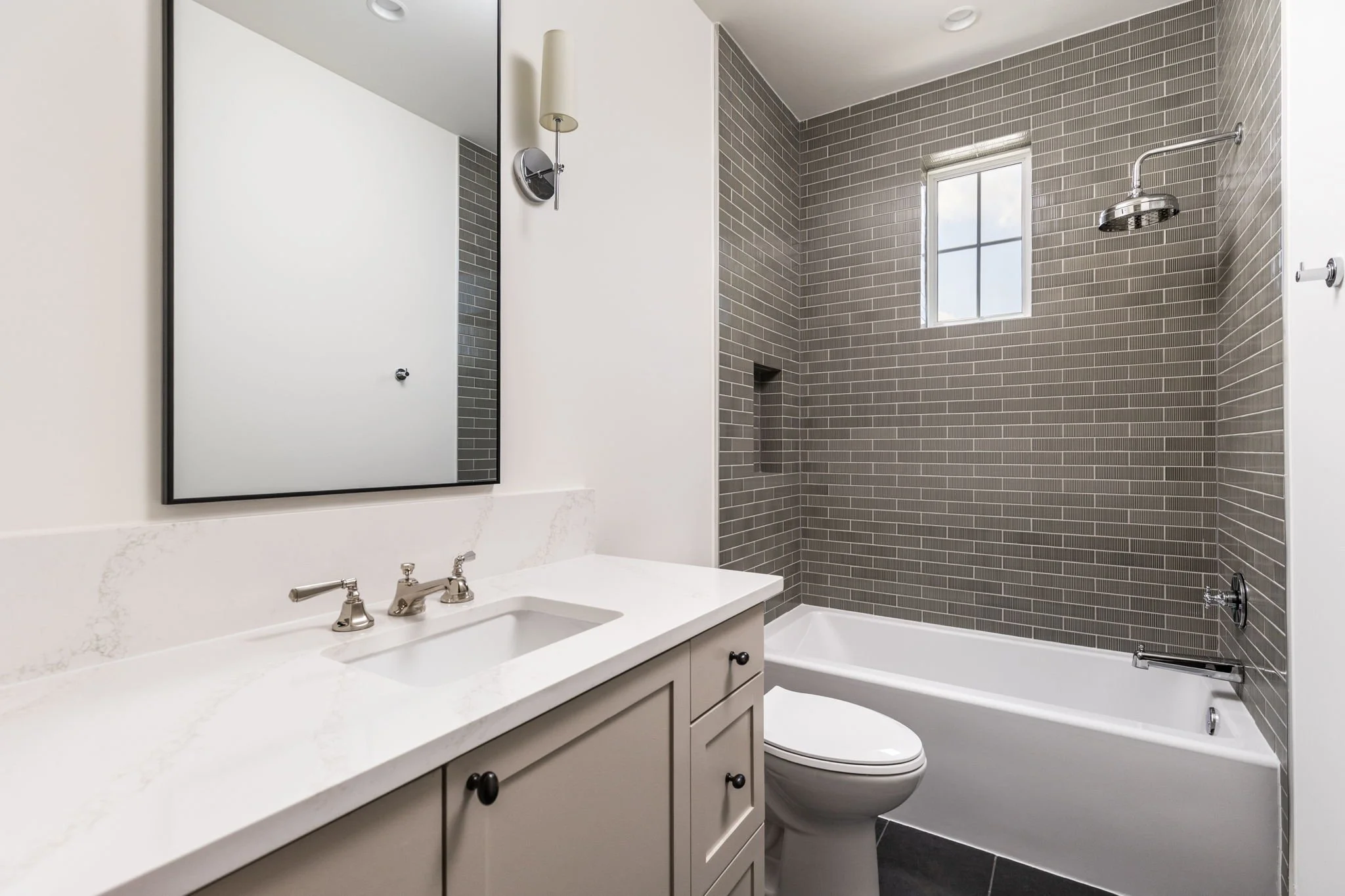 Modern bathroom with gray tile walls, white vanity with marble countertop, large mirror, and a bathtub with a window and showerhead.