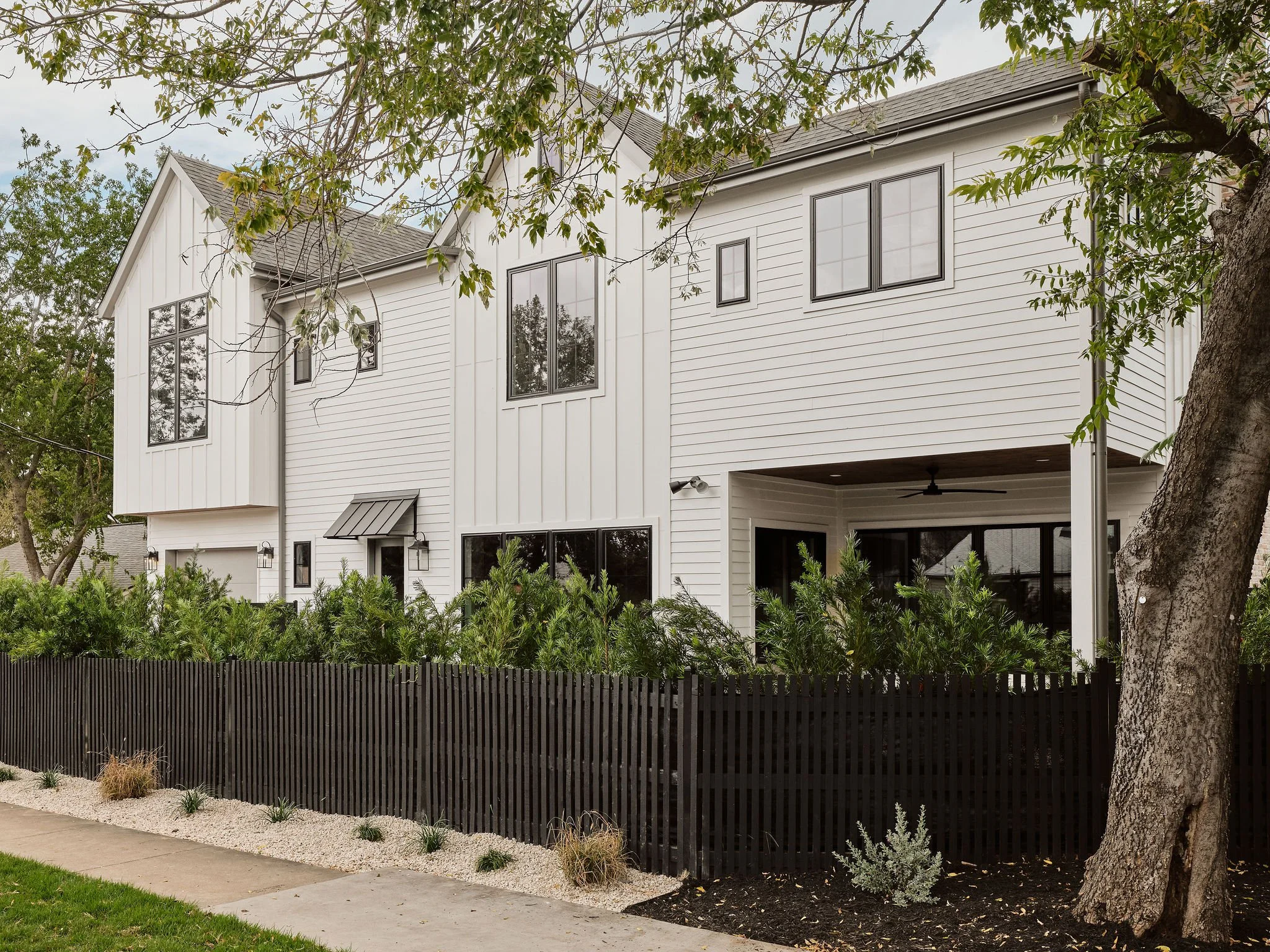 Modern white multi-story house with black-framed windows, black fence, greenery, and a sidewalk in front.