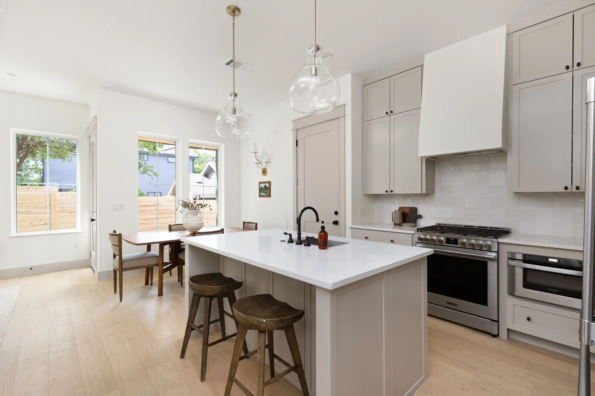 Modern kitchen with white cabinets, a large island with a white countertop, a stainless steel gas oven, and two glass pendant lights hanging from the ceiling. Dining area with a wooden table and chairs, windows with a view of neighboring houses and a