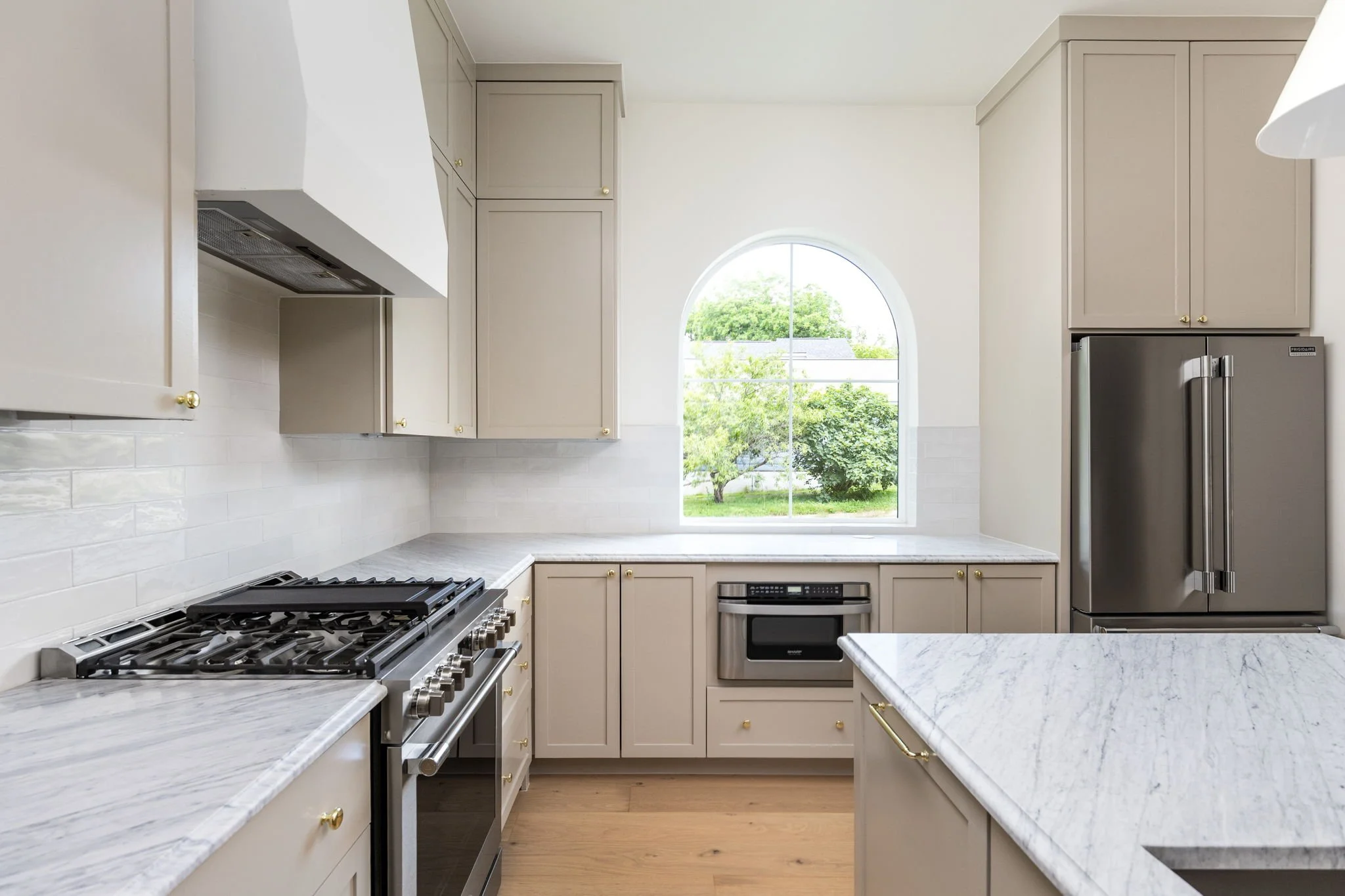 Kitchen with beige cabinets, marble countertops, a large arched window showing green trees, a stainless steel refrigerator, and a gas stove.