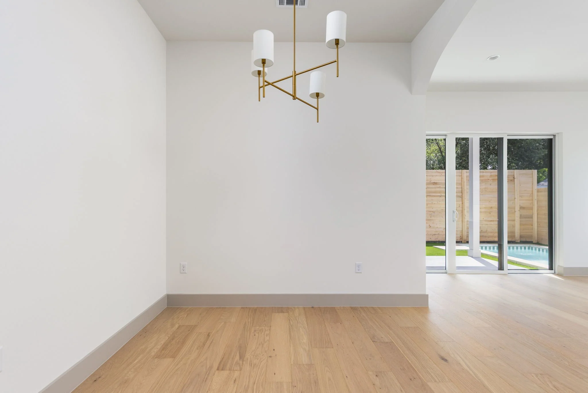Empty room with white walls, wooden flooring, modern white and gold chandelier, large sliding glass door leading to backyard with swimming pool and wooden fence.
