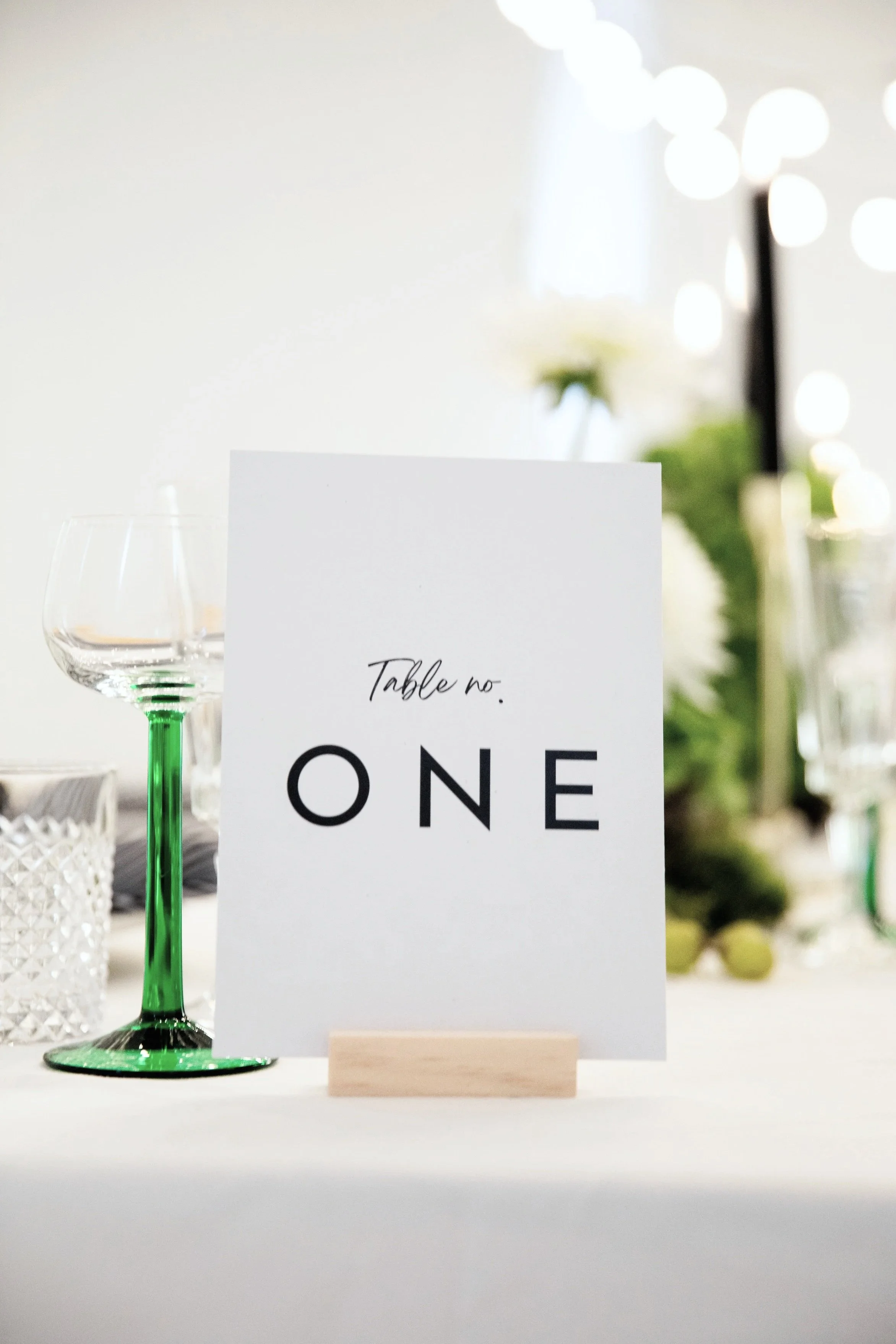 Table setting with a white tablecloth, glasses with green stems, a white card with "Table no. ONE" written on it, and a blurred background with string lights and floral arrangements.