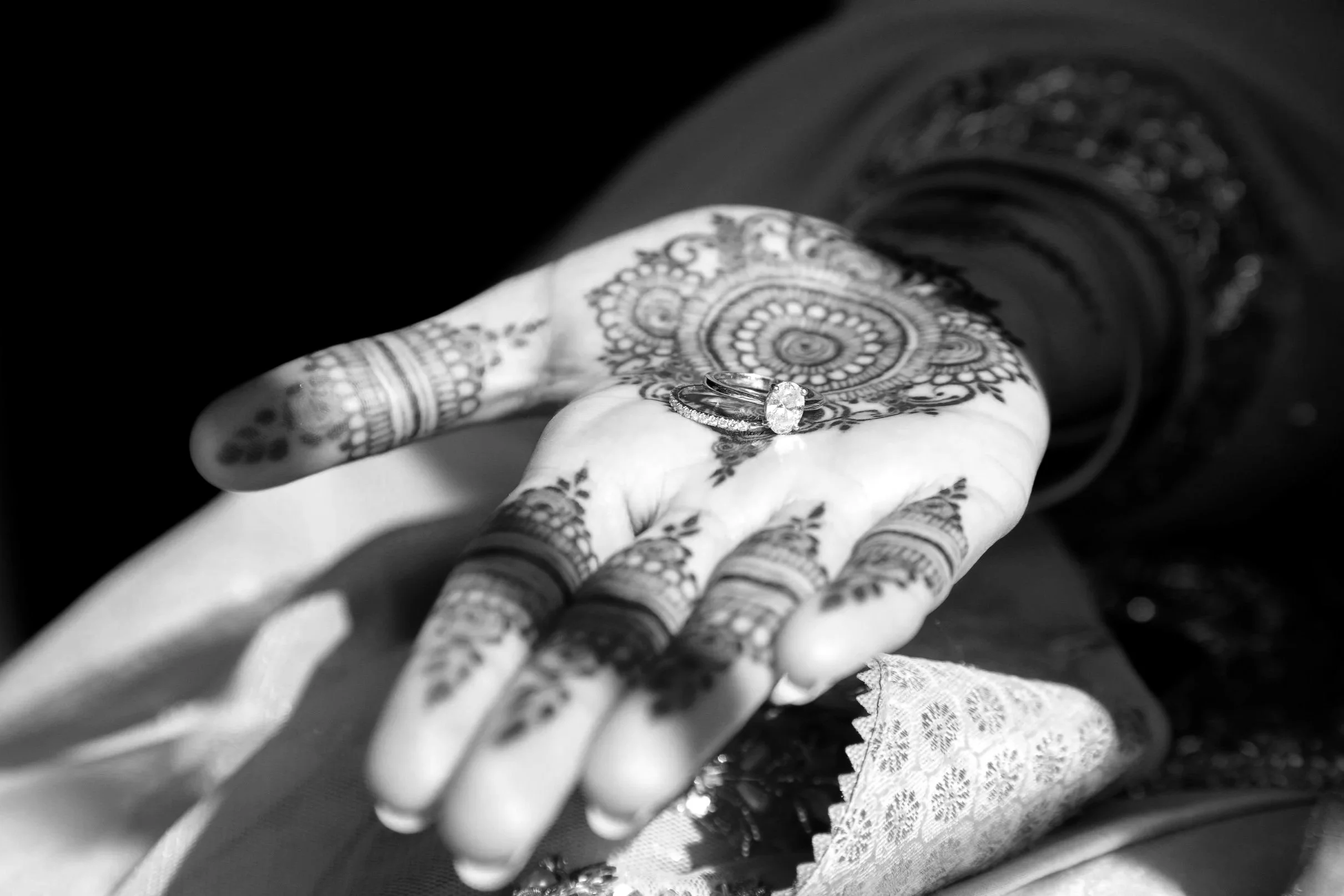Close-up of a hand decorated with orange henna designs, holding two rings: a large engagement ring with a central diamond and a wedding band with smaller diamonds.