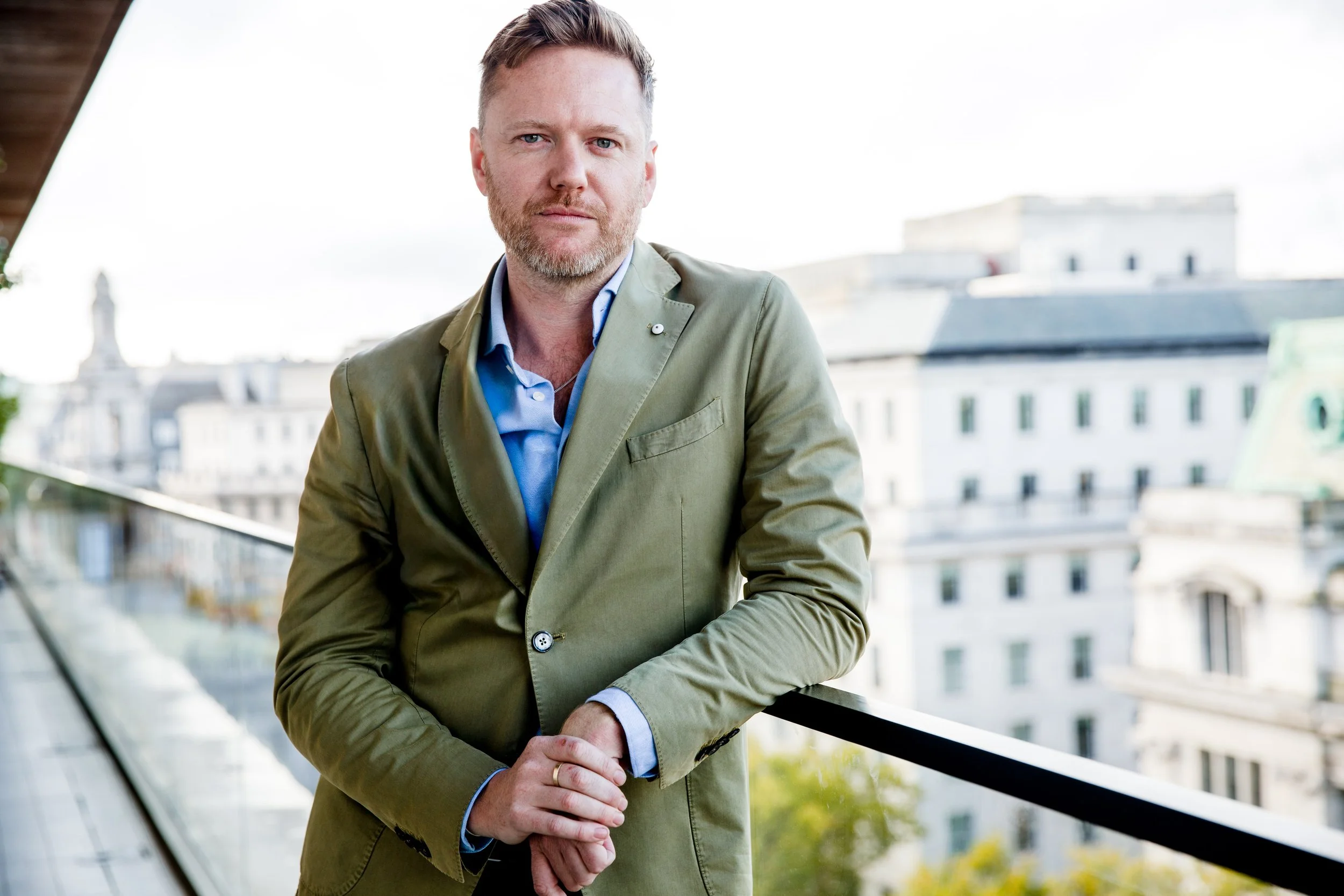 A man with light hair, beard, and blue eyes wearing a green blazer and light blue shirt, leaning on a railing outdoors with city buildings in the background.