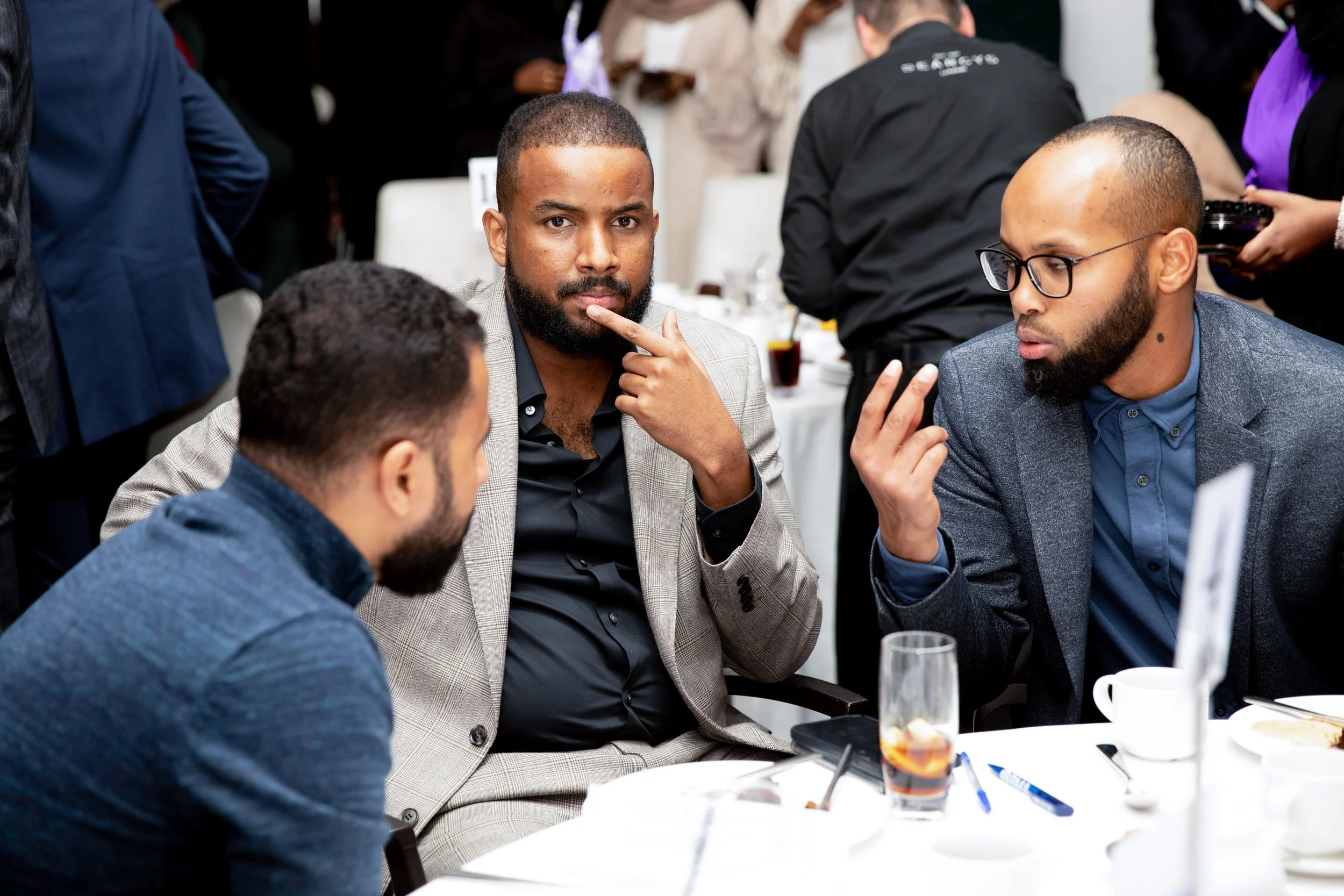 Three men engaged in a conversation at a table during an event, with drinks and food on the table and people in the background.