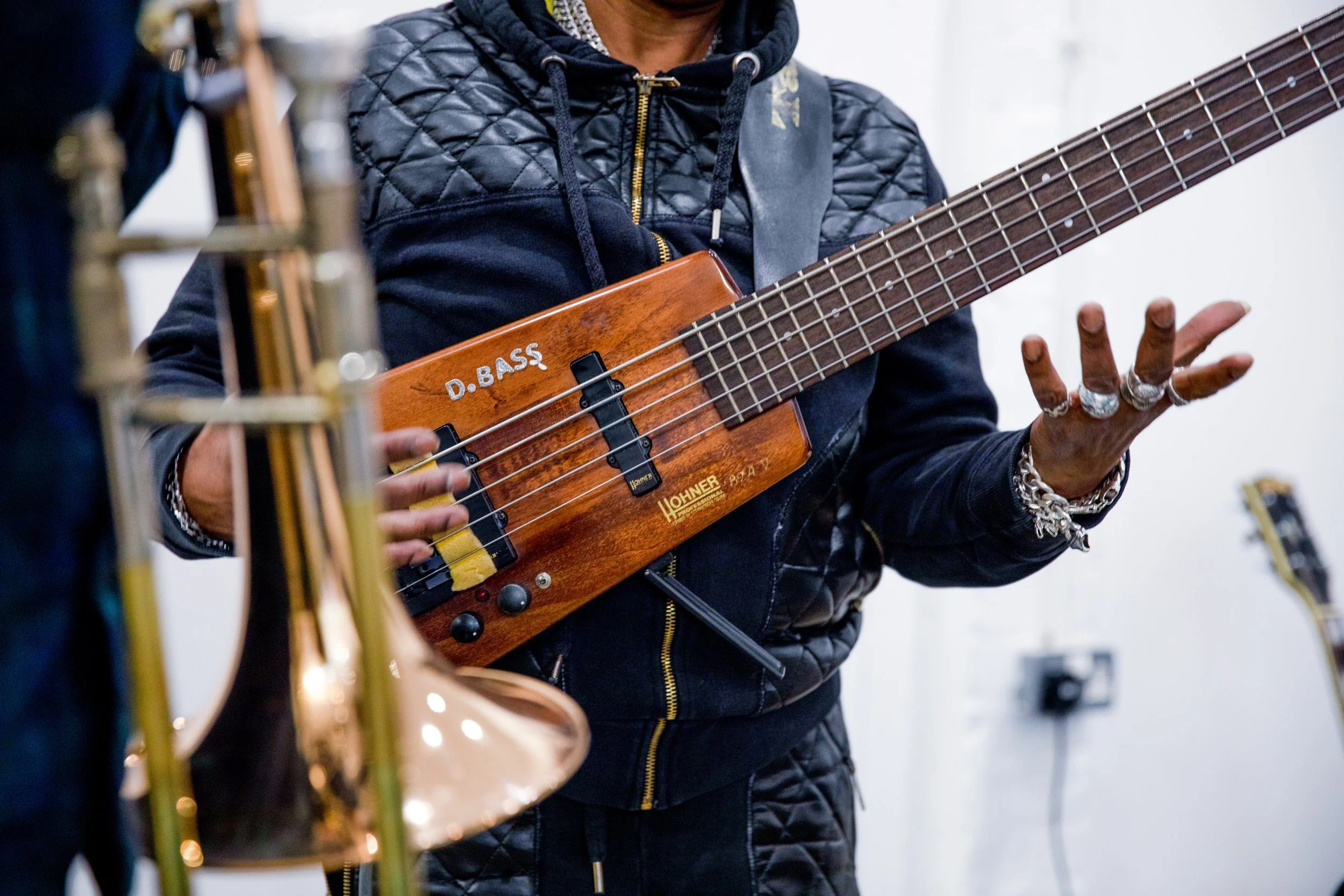 Person wearing a quilted black jacket and silver rings playing a wooden bass guitar labeled 'D. BASS' in a music rehearsal space.