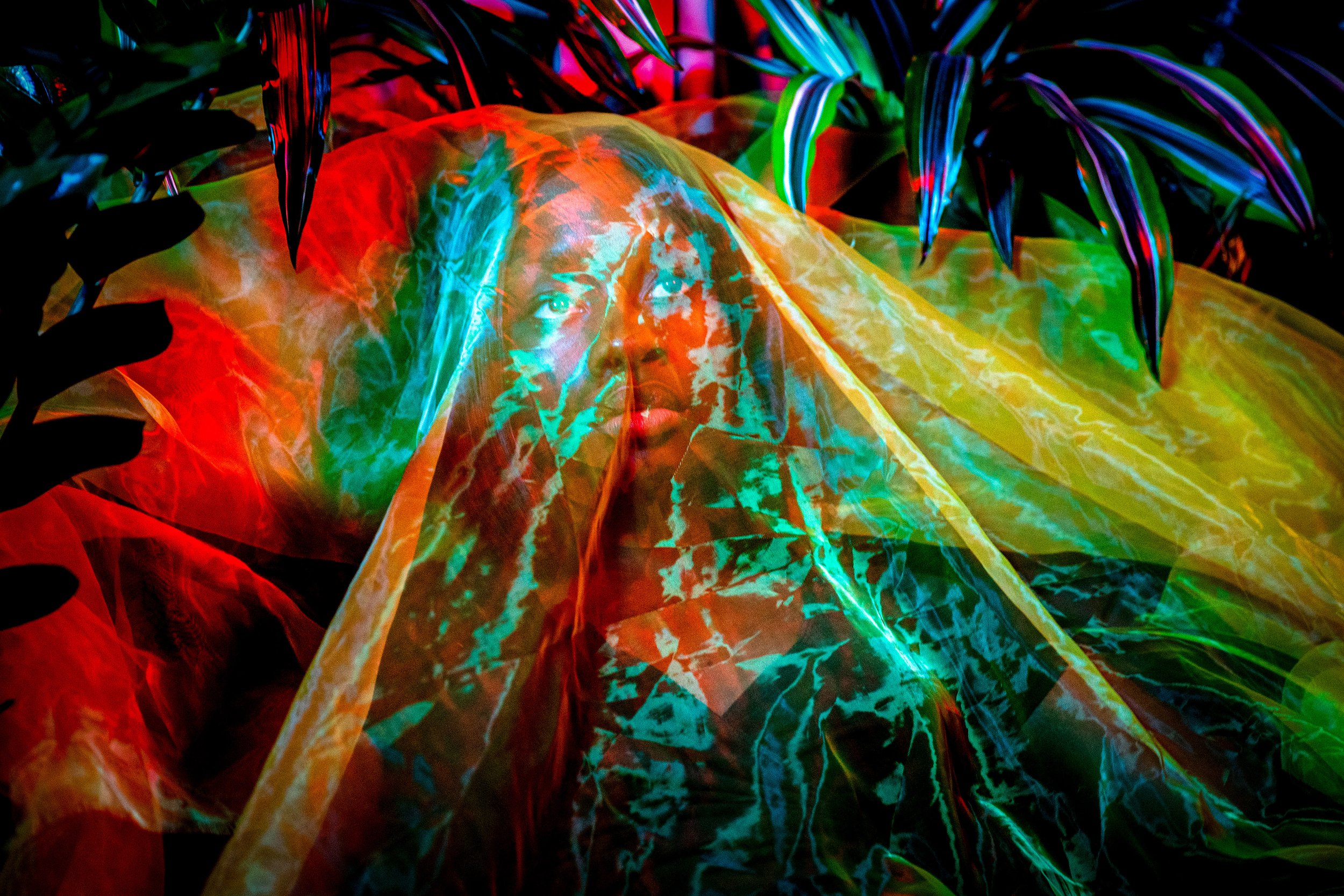 A person with a light complexioned face looking upwards is behind a colorful, neon-illuminated fabric or surface, with vibrant green, red, blue, and yellow hues creating abstract patterns.