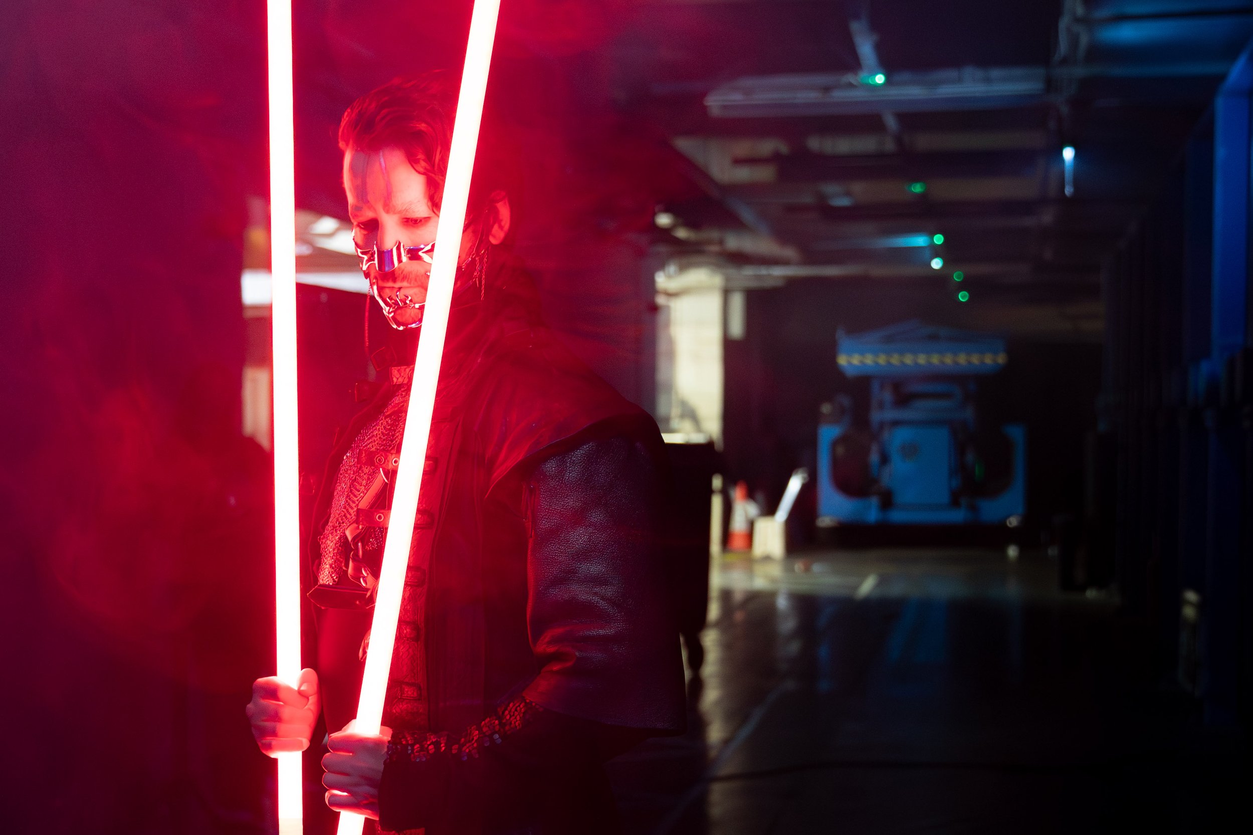 Person dressed in dark clothing holding two glowing red light rods in a dark indoor setting, with industrial equipment and ceiling lights in the background.