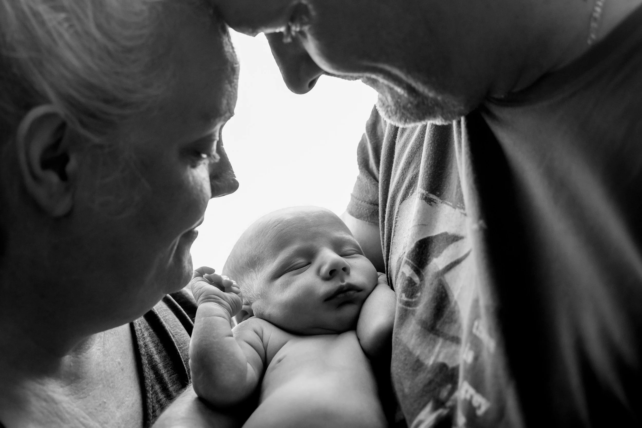 A close-up black and white photograph of a family, showing a man, woman, and a sleeping baby. The man and woman are leaning closely towards each other, with their foreheads touching, and the woman is holding the baby gently.