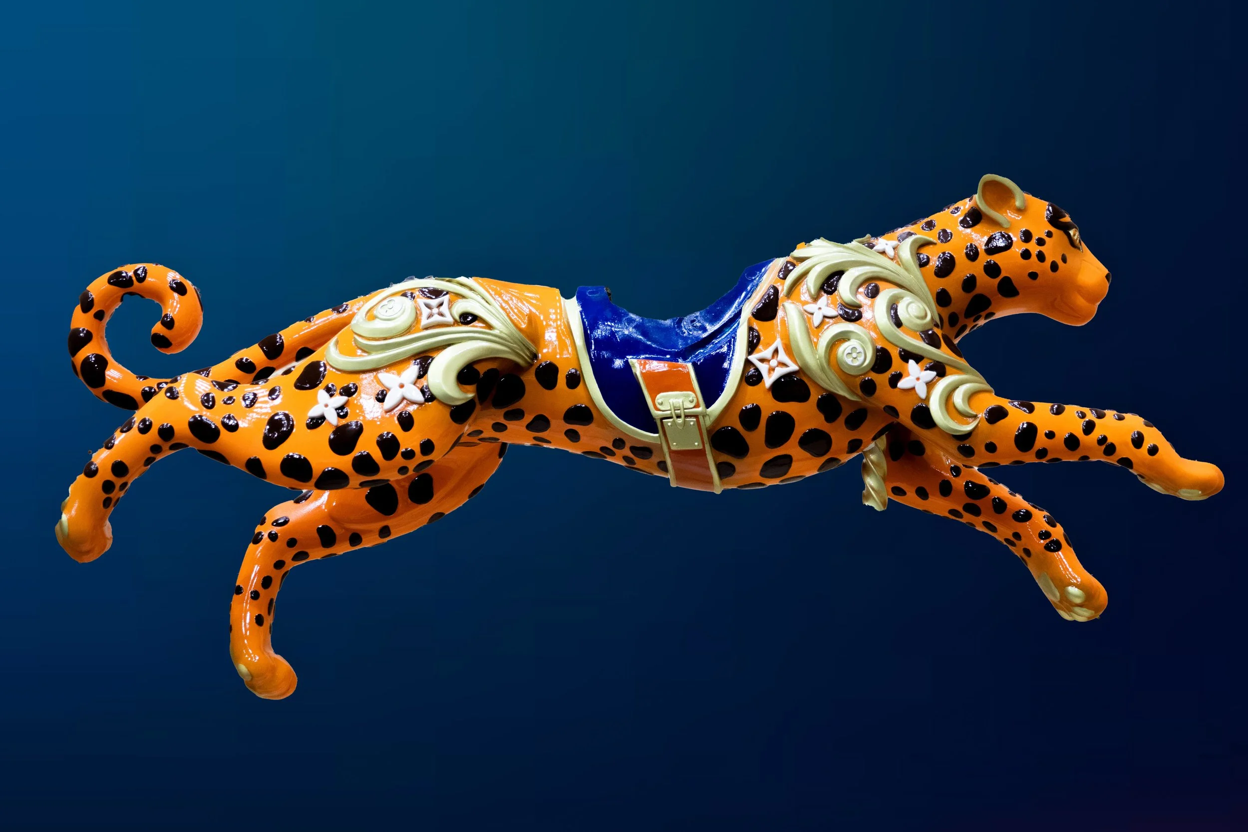 A decorative sculpture of a cheetah with orange and black spots, adorned with intricate floral and geometric patterns, placed against a solid blue background.