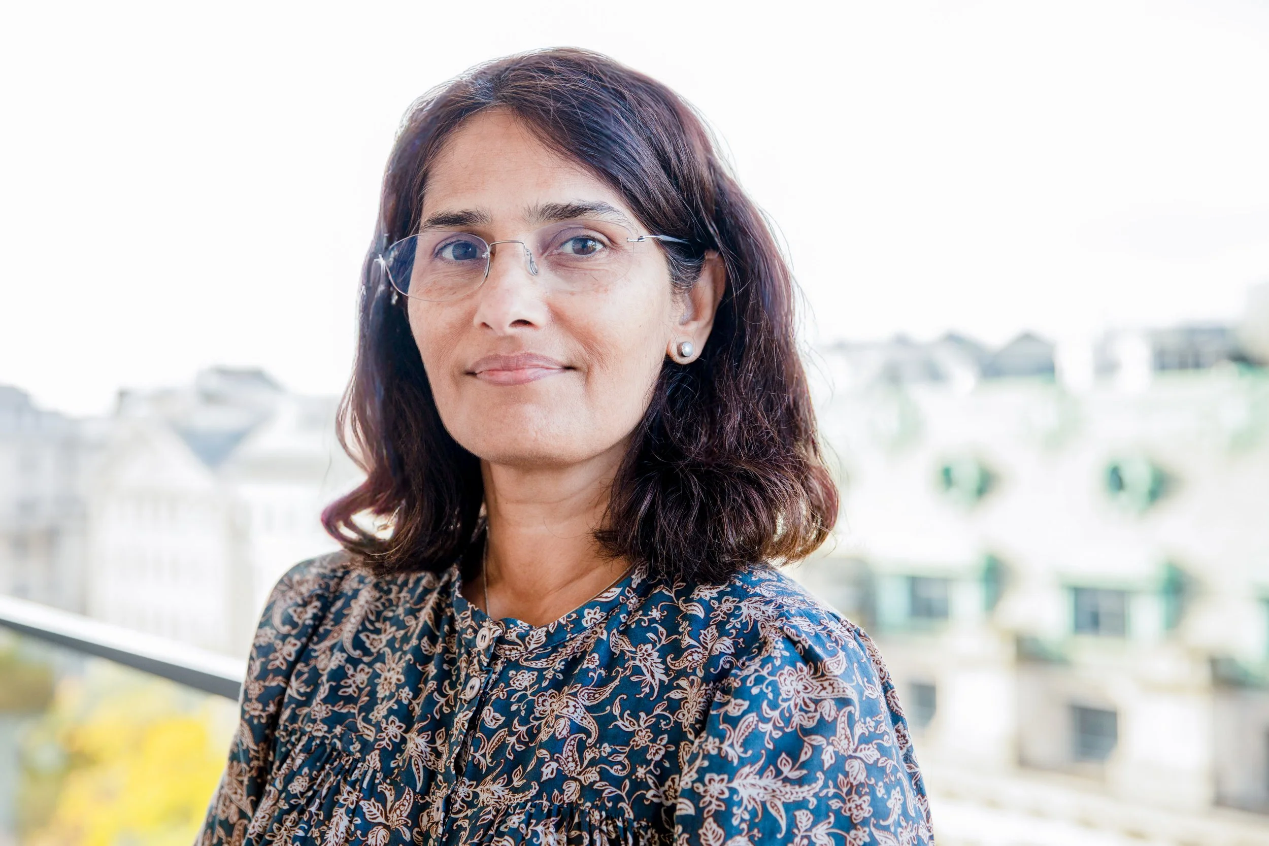 A woman with shoulder-length brown hair and glasses, wearing a blue floral blouse, standing outdoors with a blurred background of buildings and a body of water.
