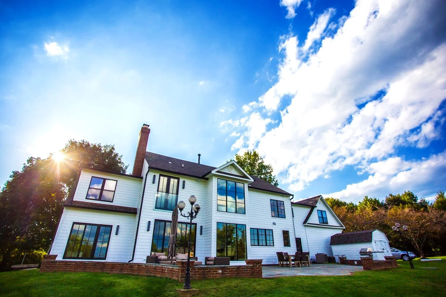 Large white house with large windows and brick trim, set against a blue sky with clouds and the sun shining. The house has a sloped roof and outdoor seating on a patio, surrounded by grass and trees.