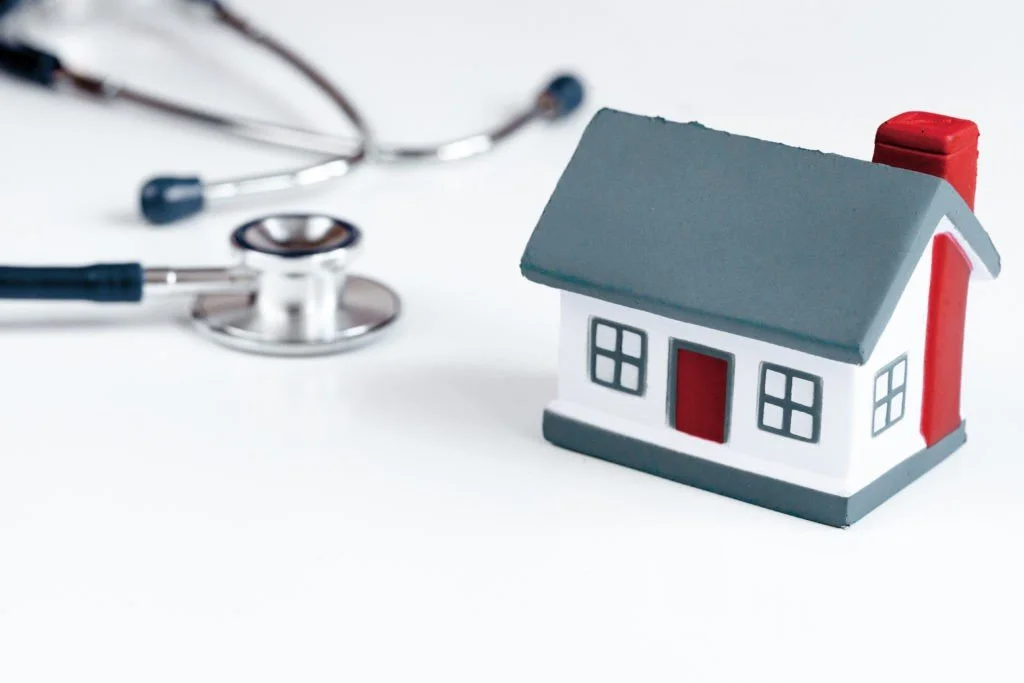 A small model house with red and gray accents next to a stethoscope on a white surface.