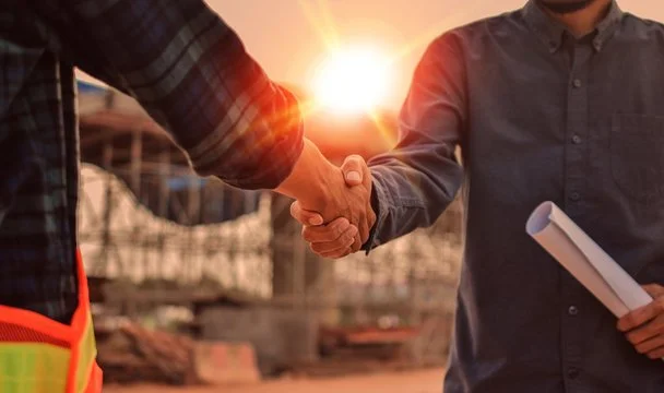Two men shaking hands at a construction site with the sun setting in the background.