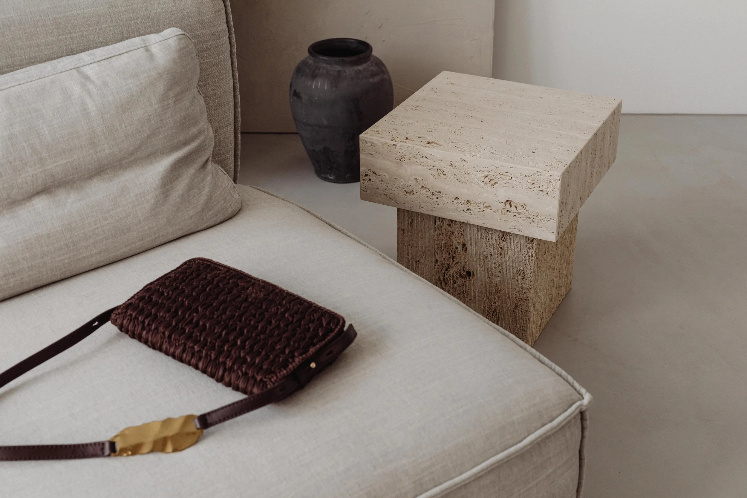 Ein Sofa mit beige Kissen, eine braune handgefertigte Tasche mit Gürtel auf der Sitzfläche, ein schwarzer Vasen und ein quadratischer Beistelltisch aus hellem Stein und Holz im Hintergrund.