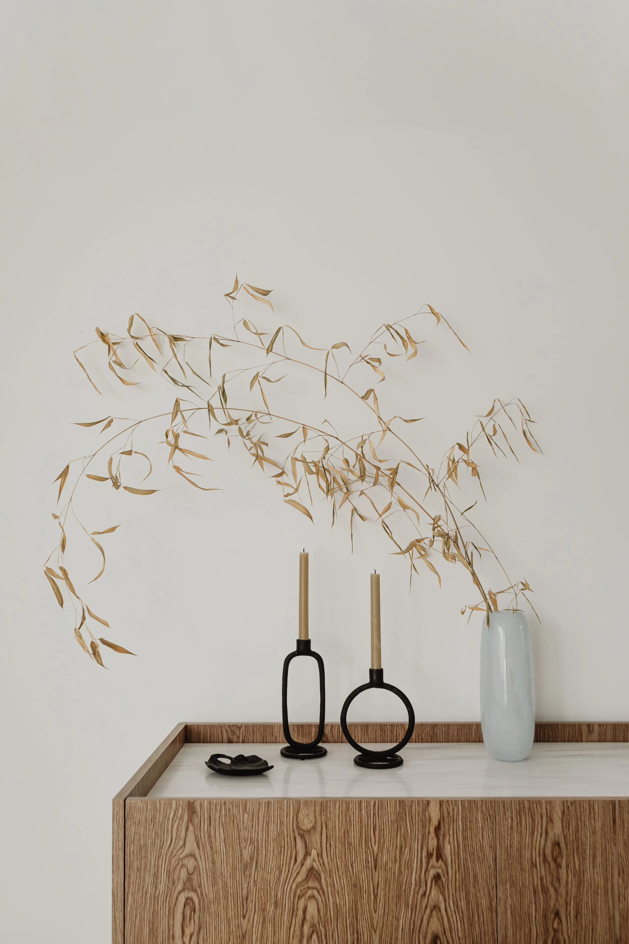 Eine Holzkonsole mit zwei schwarzen Kerzenhaltern, einer weißen Vase mit getrockneten Ästen und einem kleinen schwarzen Gegenstand.