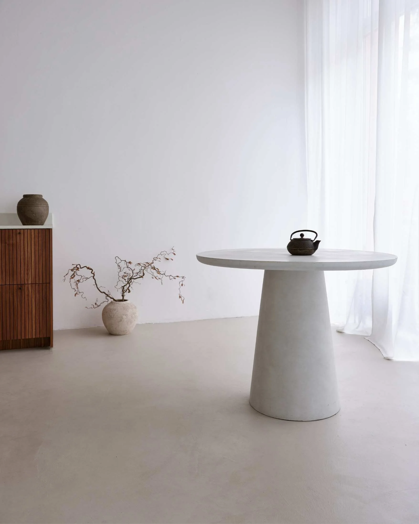 minimalistischer Raum mit weißen Wänden und Boden, einem runden weißen Tisch mit einem schwarzen Teekessel darauf, und einer weißen Vase mit verzweigten Ästen, sowie einem kleinen braunen Keramikvase auf einem Holzsideboard im Hintergrund