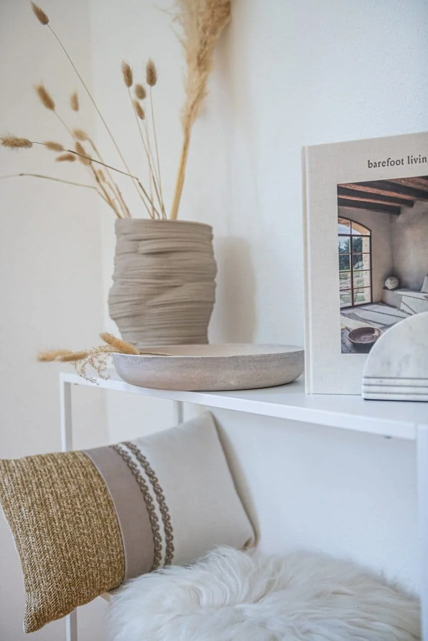 Inneneinrichtung mit einer weißen Konsole, einem dekorativen Krug mit getrockneten Gräsern, einer neuwertigen, künstlerisch gestalteten Schale und einem Buch mit großem Bild eines modern eingerichteten Wohnzimmers.