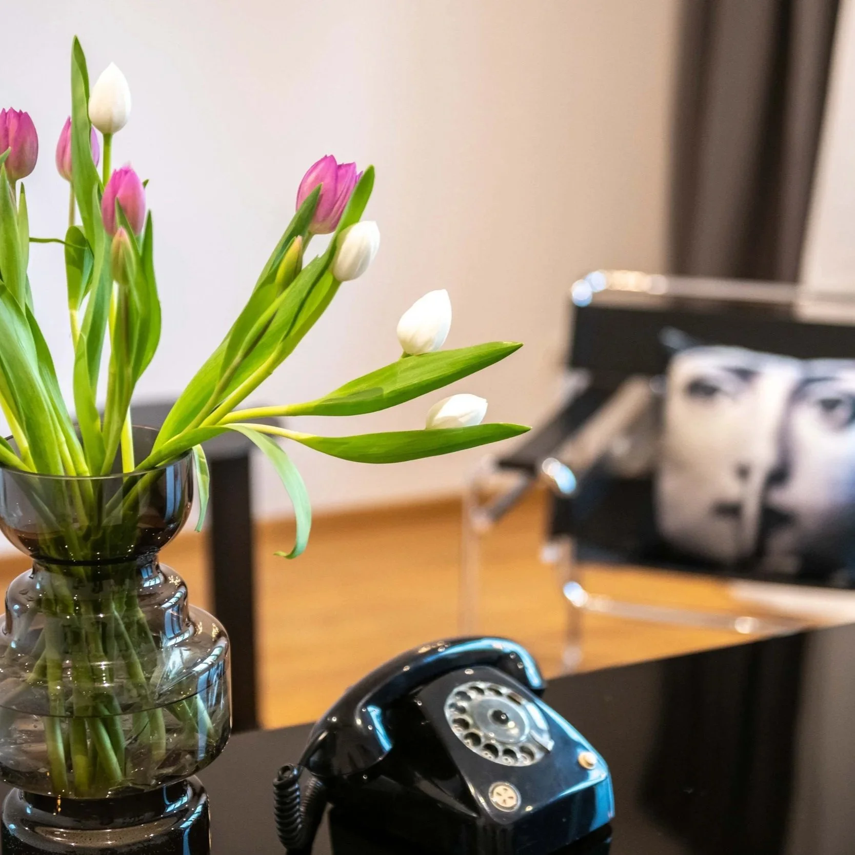 Vase mit weißen und rosa Tulpen auf einem schwarzen Tisch, im Hintergrund ein altes schwarzes Telefon und ein Kissen mit einem Gesicht drinnen