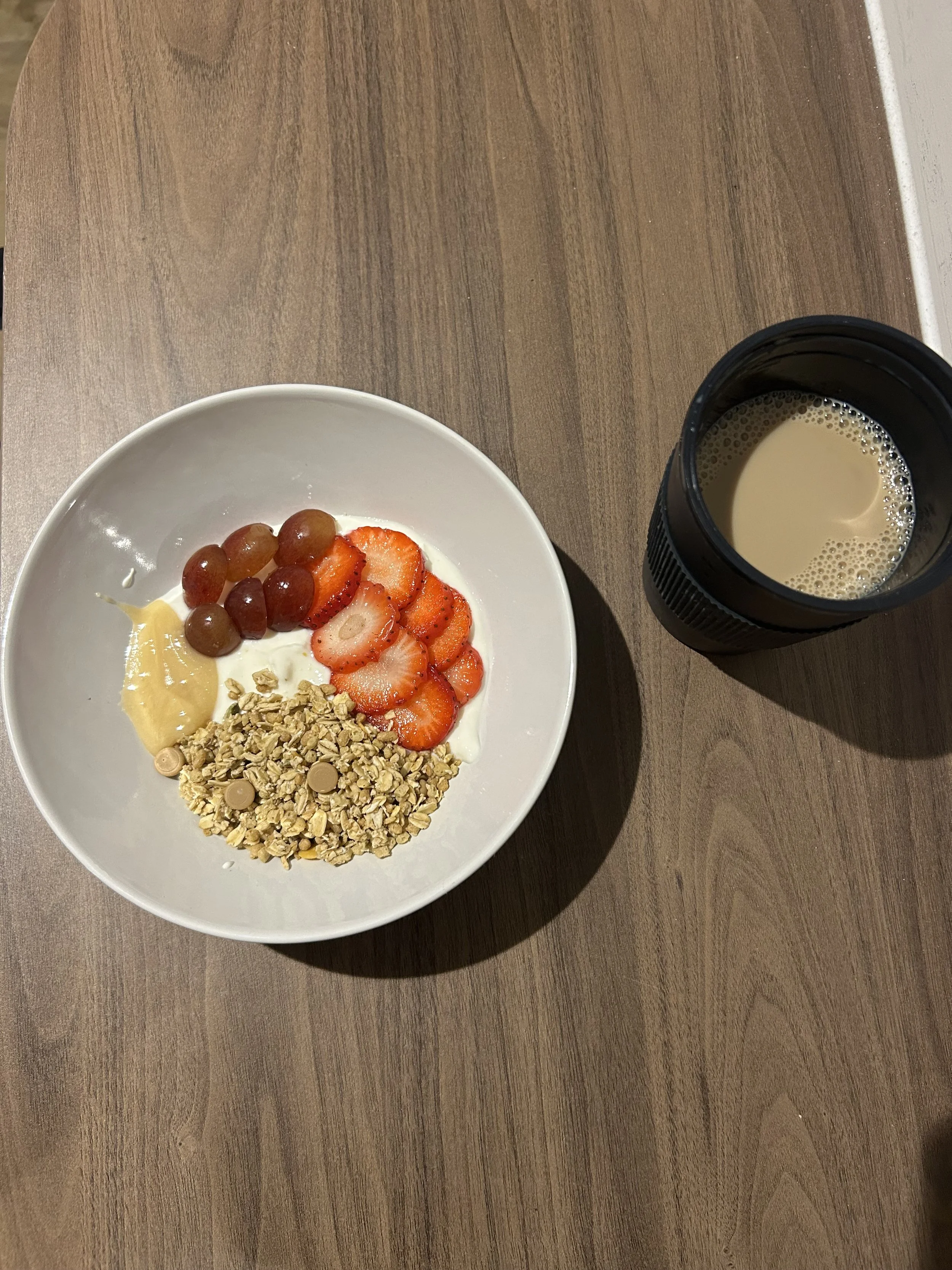 A white bowl containing grapes, sliced strawberries, granola with chocolate chips, and a dollop of honey, with a cup of coffee or tea next to it on a wooden surface. This is a nutrition focused meal that coaches can advise to clients.