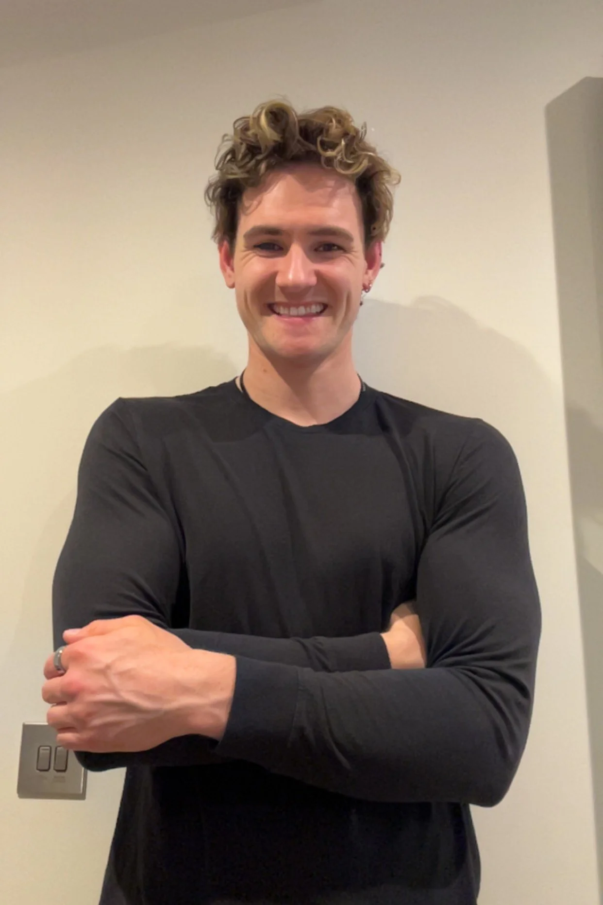 A young man with styled curly blond hair, smiling, wearing a black long-sleeve shirt, standing with arms crossed against a plain wall