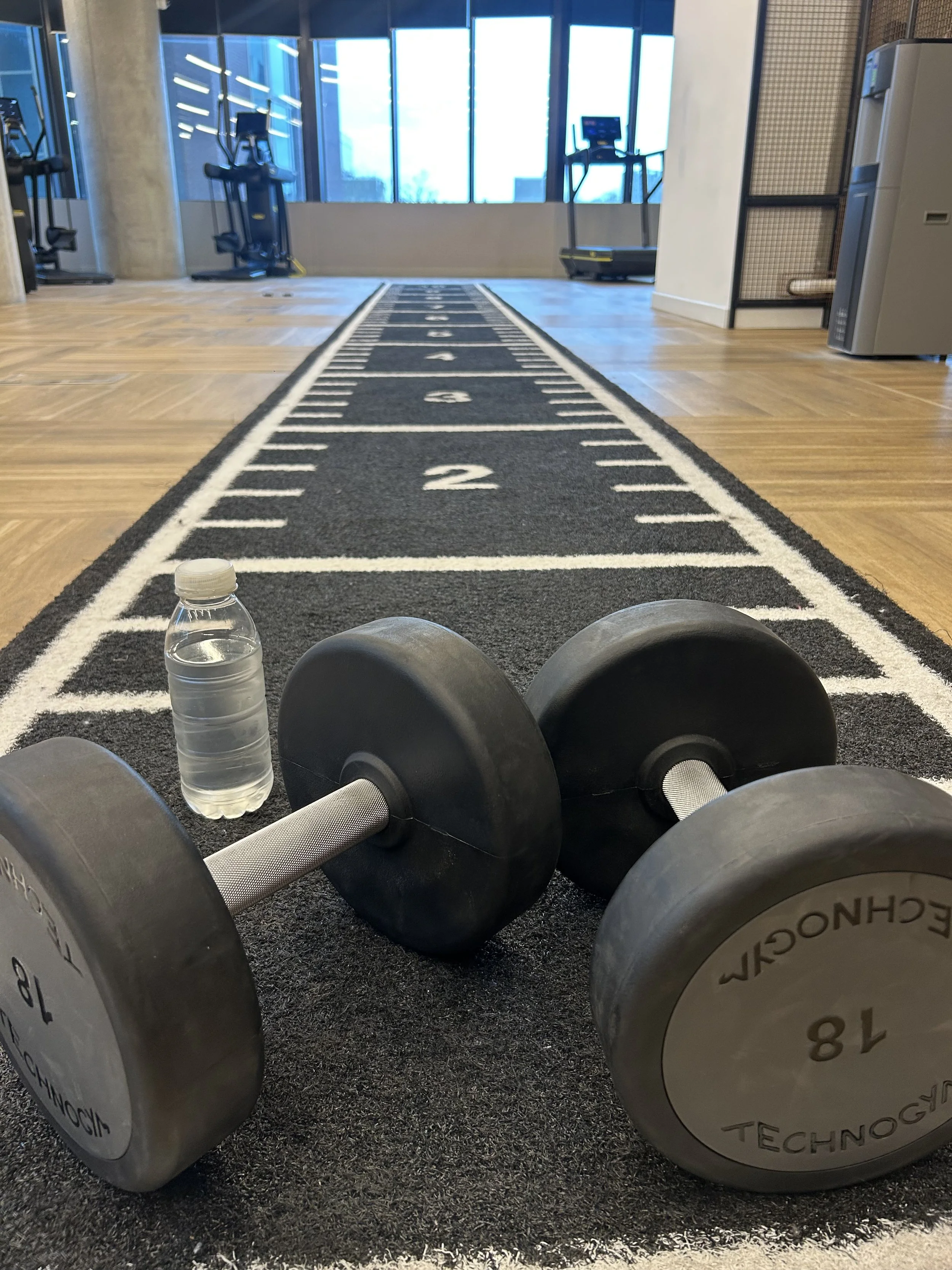 Dumbbell and water bottle on a black running track mat inside a gym with large windows in the background. These items are to be used for personal training in the gym.