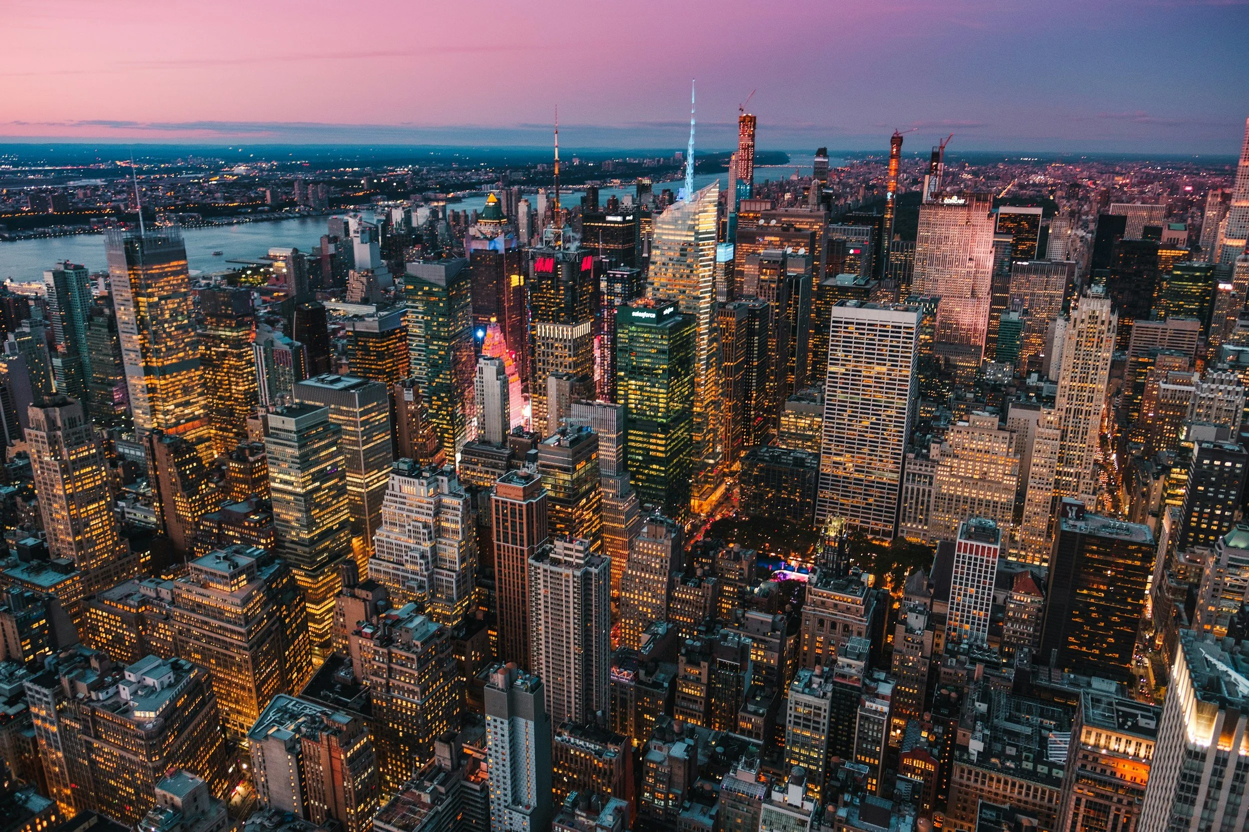 Aerial view of a city skyline at sunset with tall skyscrapers and illuminated buildings in New York City, overlooking the Hudson River.