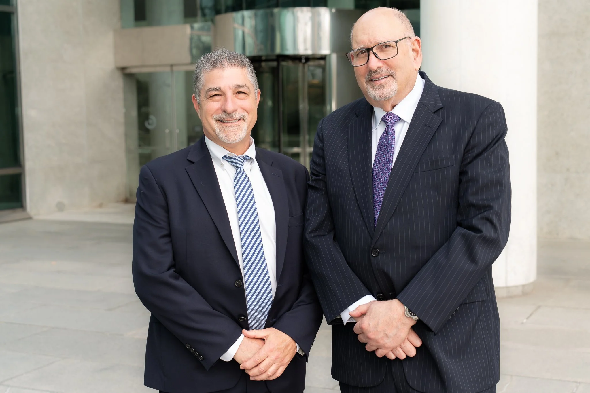 Two middle-aged men in business suits standing outside in front of a modern building, smiling at the camera.