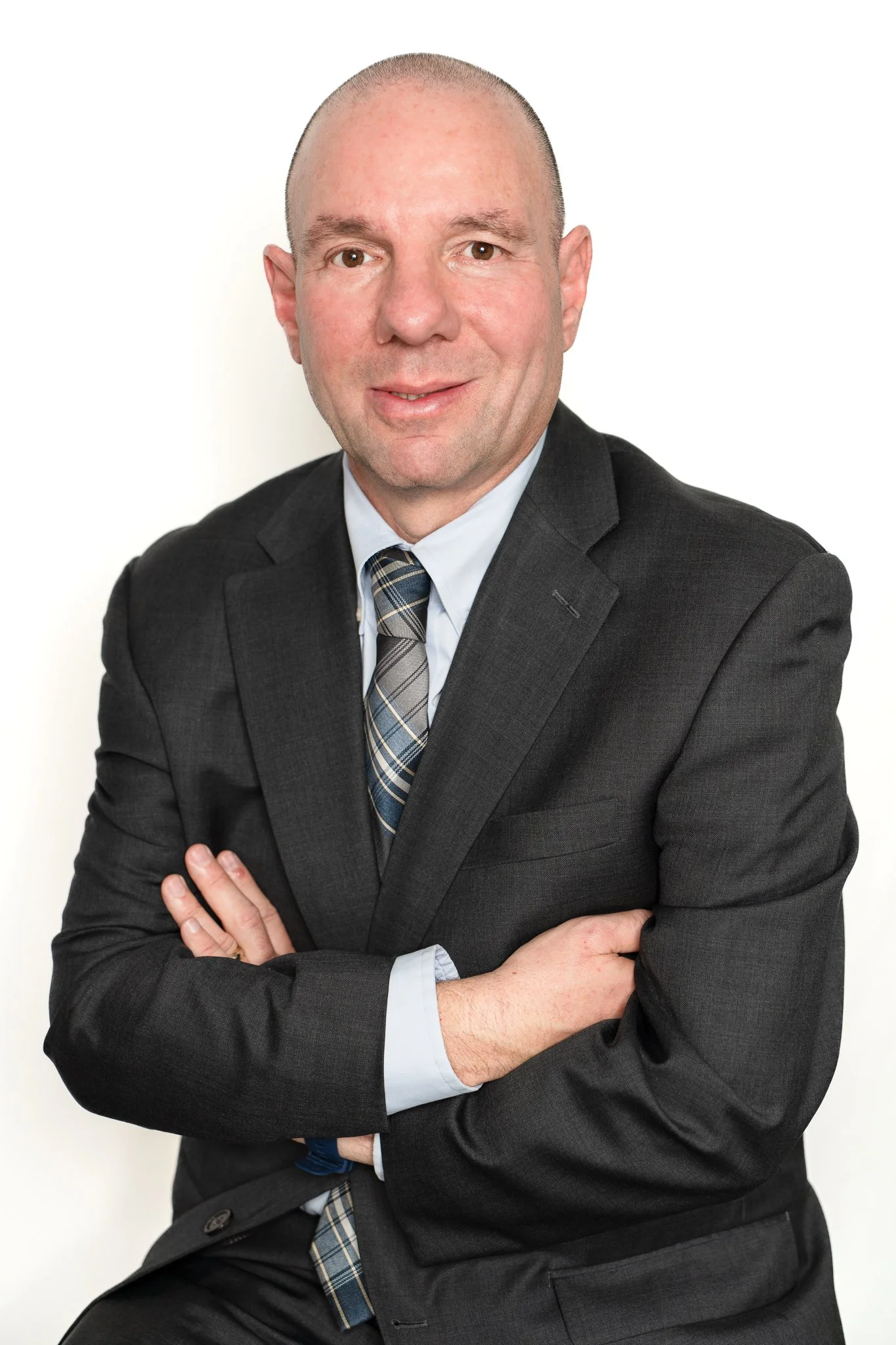A man in a dark suit, light blue shirt, and plaid tie, with arms crossed, posing against a white background.