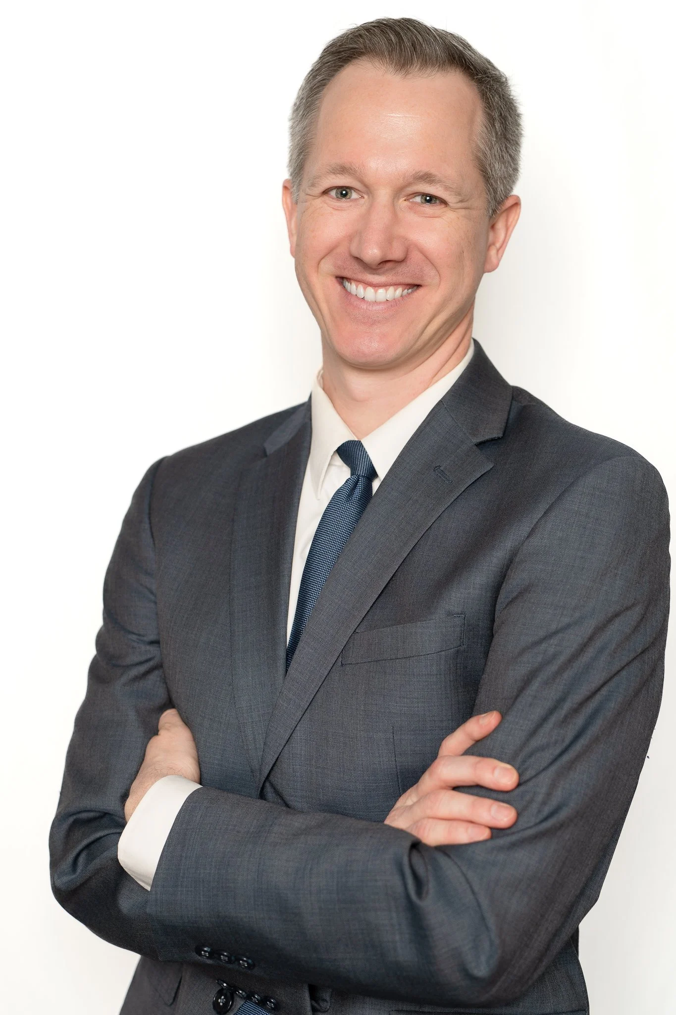 A smiling man with short gray hair in a dark gray suit, white shirt, and navy tie, crossing his arms against a white background.